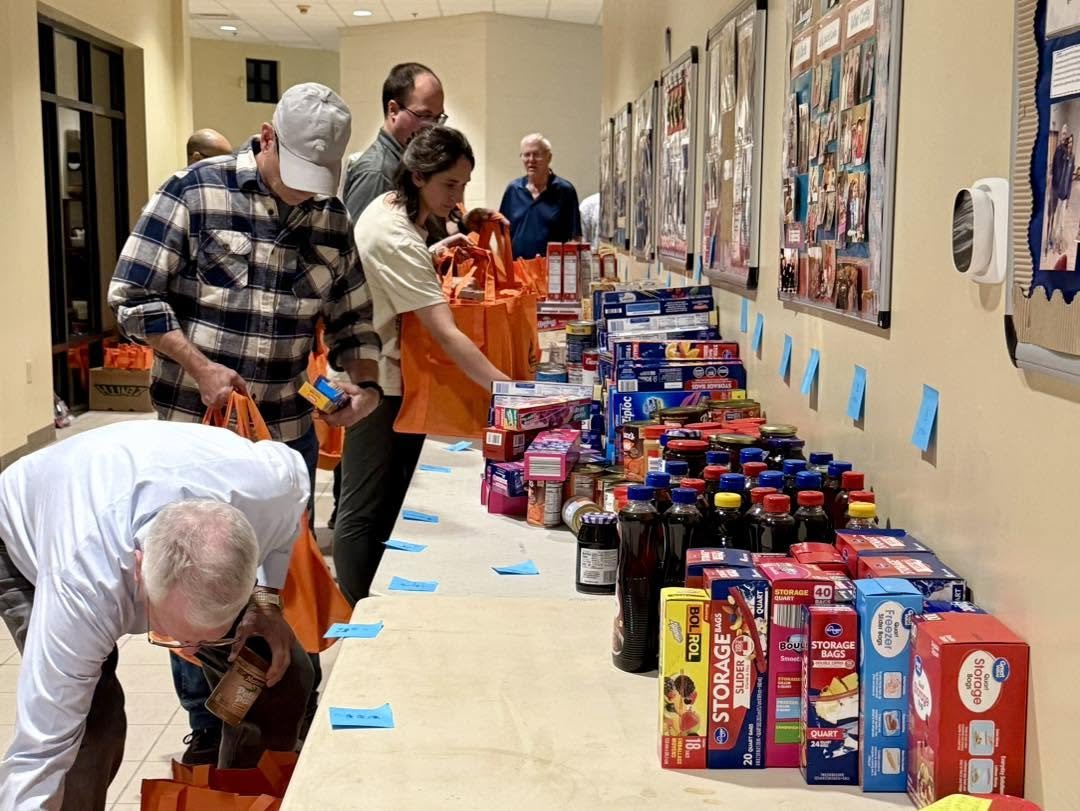 People shopping for groceries in a line at a donation event, with non-perishable food items and supplies on a long table.