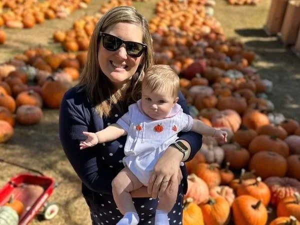A woman holding a baby girl at a pumpkin patch with numerous pumpkins on the ground.