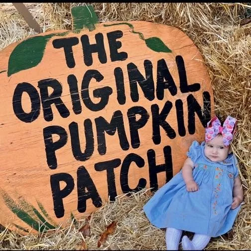 A large orange pumpkin-shaped sign with green leaves and the text "The Original Pumpkin Patch" in black letters, with a small girl in a blue dress and pink bow sitting next to it in straw.