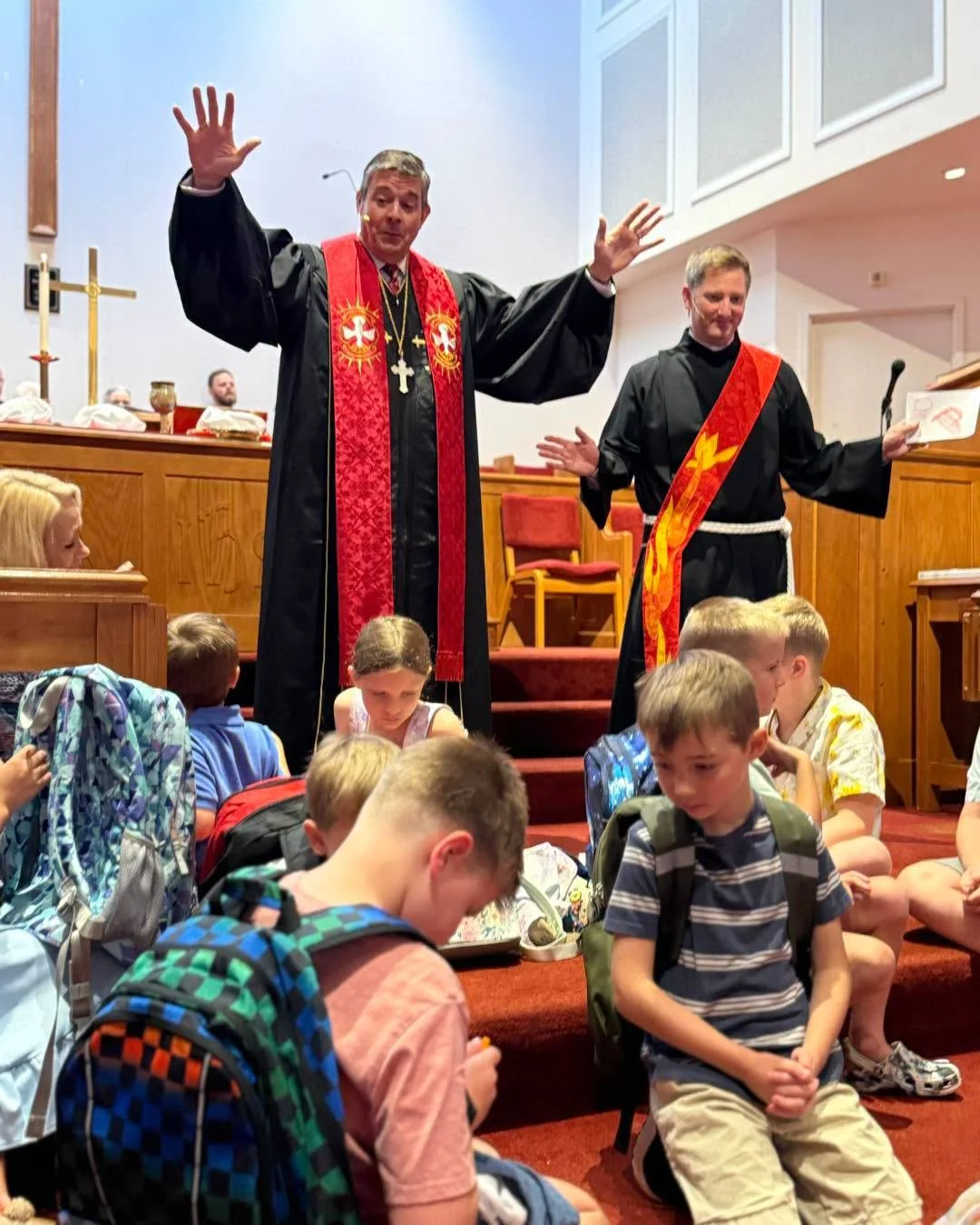 A religious service with two clergy members dressed in ceremonial robes, standing on a platform with children sitting on the steps in front, some with backpacks, inside a church.