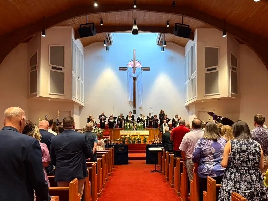 People gathered in a church with wooden pews, a large cross draped with cloth at the altar, and a choir or musicians performing at the front. The congregation is standing and facing the stage.