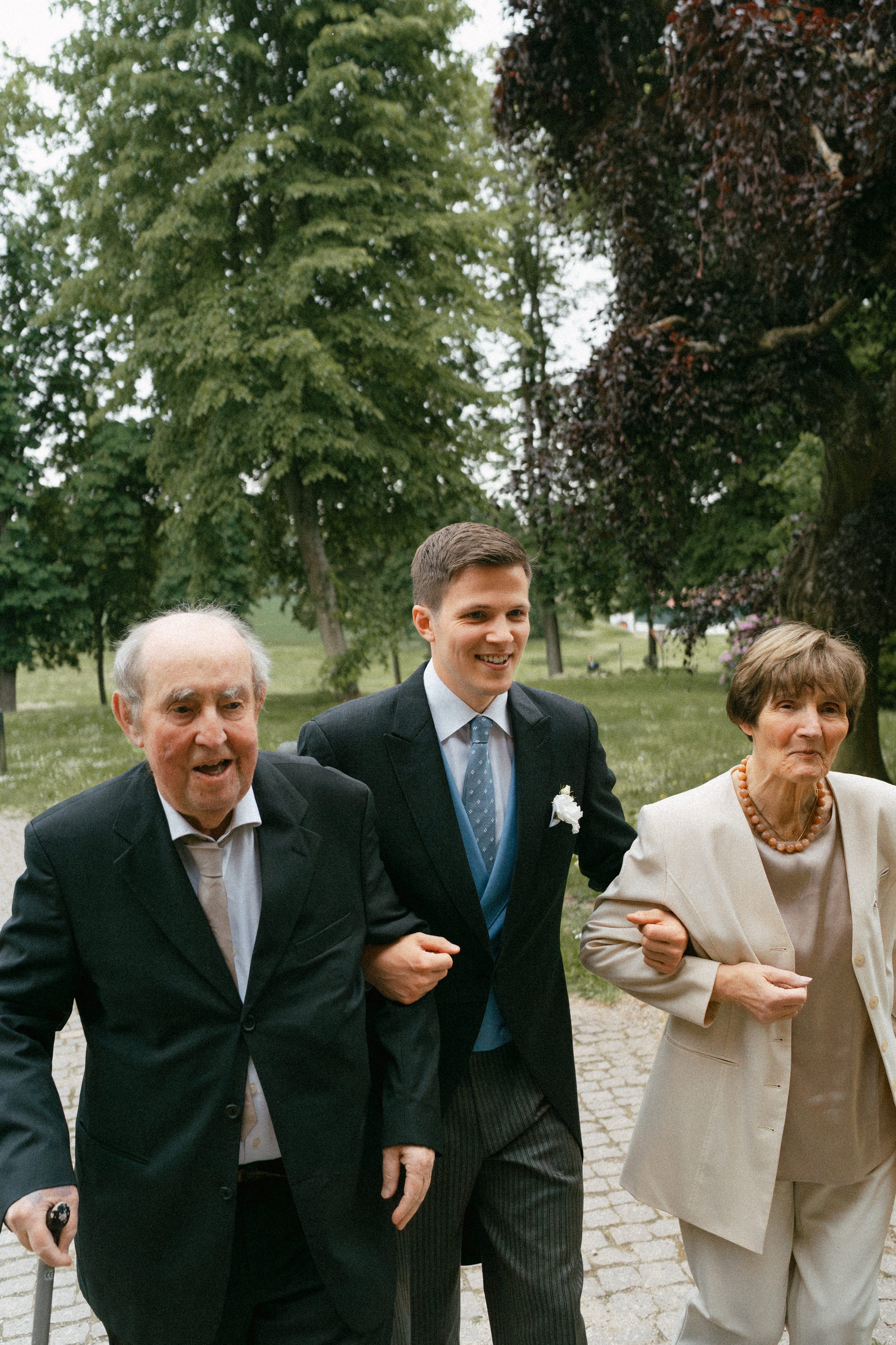 A young man wearing a suit stands between two older adults, smiling and walking together through a park. Trees and foliage are in the background.