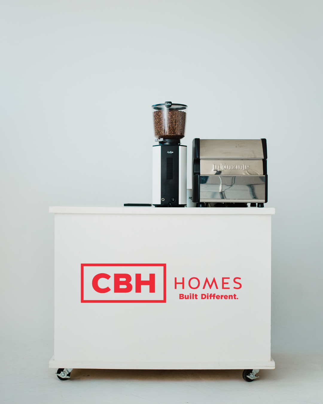 A coffee and pastry cart with a coffee grinder, an espresso machine, and a logo that reads "CBH Homes Built Different" on a white cart against a plain light background.