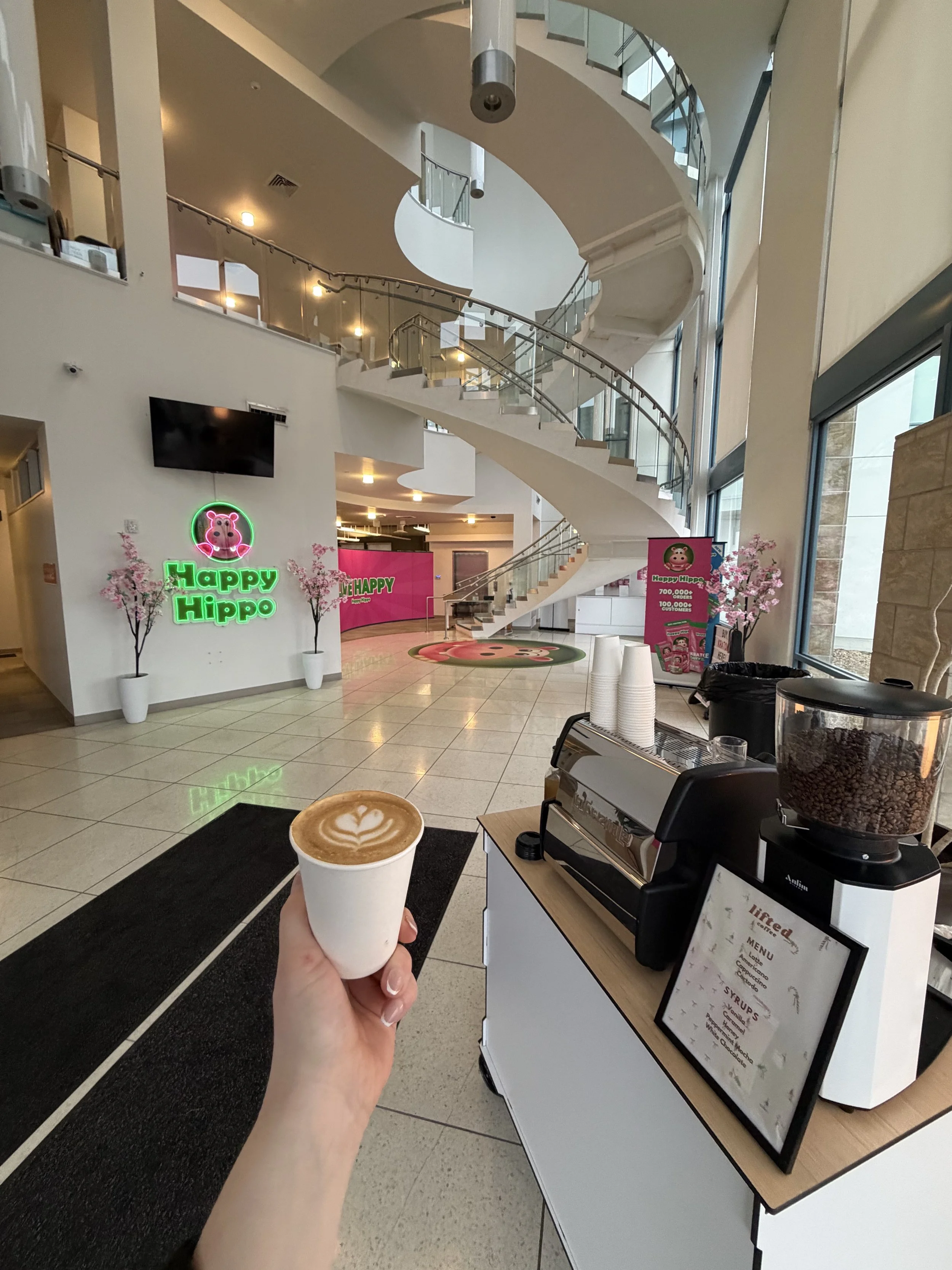 Hand holding a coffee cup with latte art in a cafe with a sign reading 'Happy Hippo' and a bright, modern interior. Lifted Coffee mobile espresso cart serving specialty coffee at private events. Custom branded bar, cups, drinks, menu and latte logo p