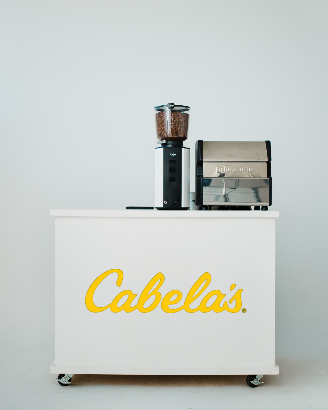 A white cart with yellow 'Cabela's' logo, topped with a coffee grinder with coffee beans and a toaster.