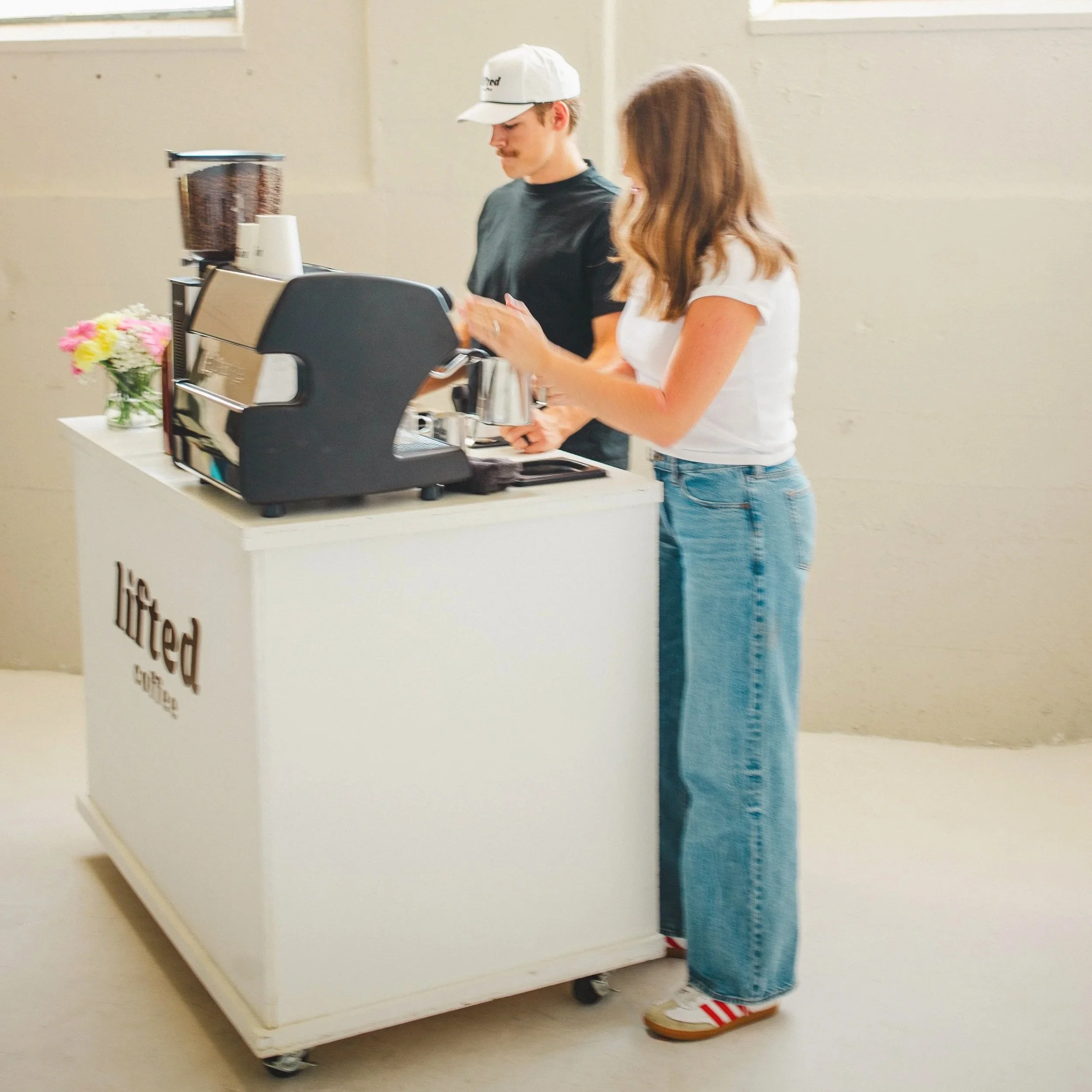 A woman and a barista making coffee at a white coffee cart with a coffee machine, flowers, and a cereal dispenser in a bright, minimal room.