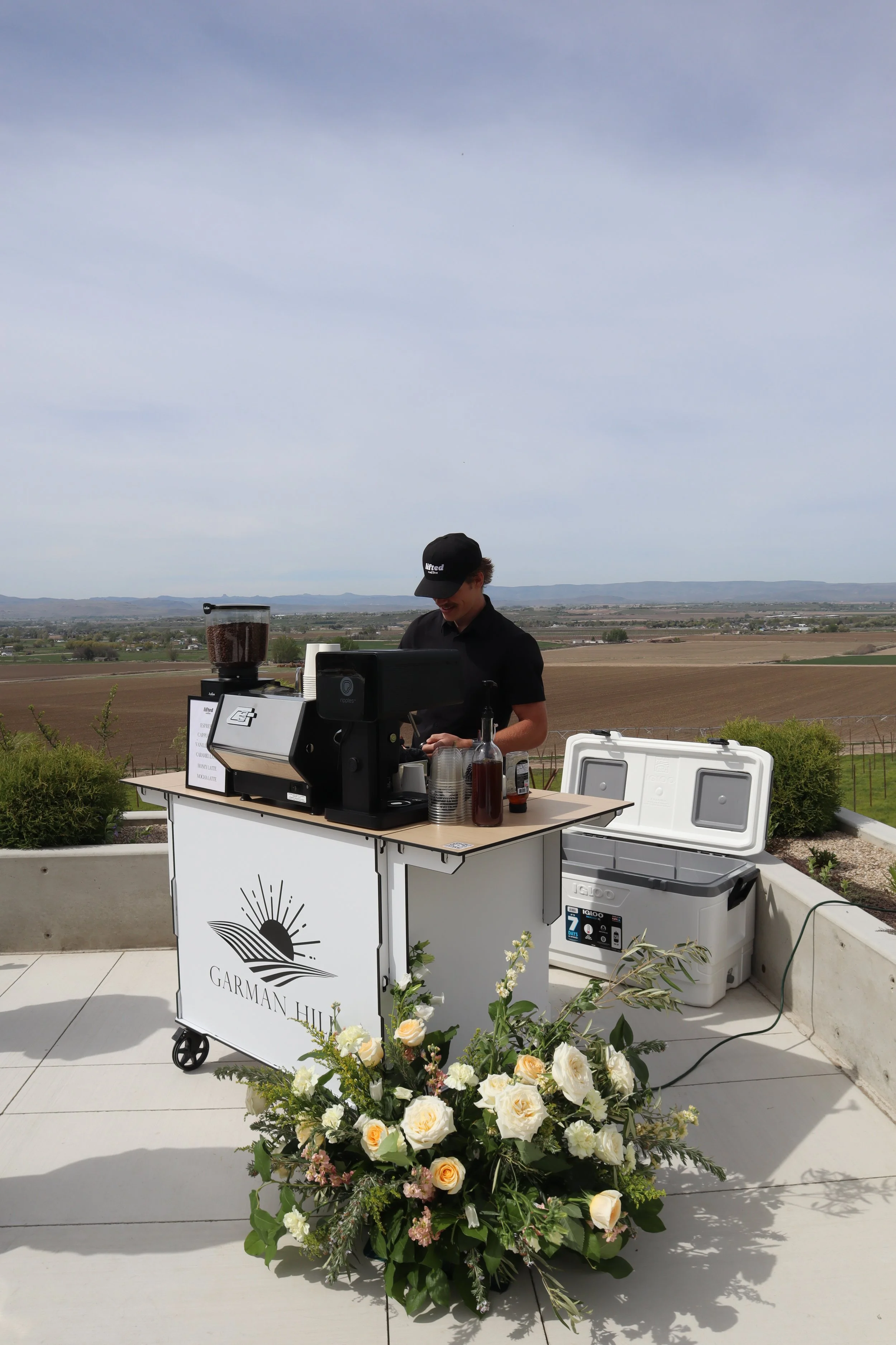 A person operating a mobile coffee cart outdoors with a vast landscape of fields and mountains in the background, and a large floral arrangement in the foreground.