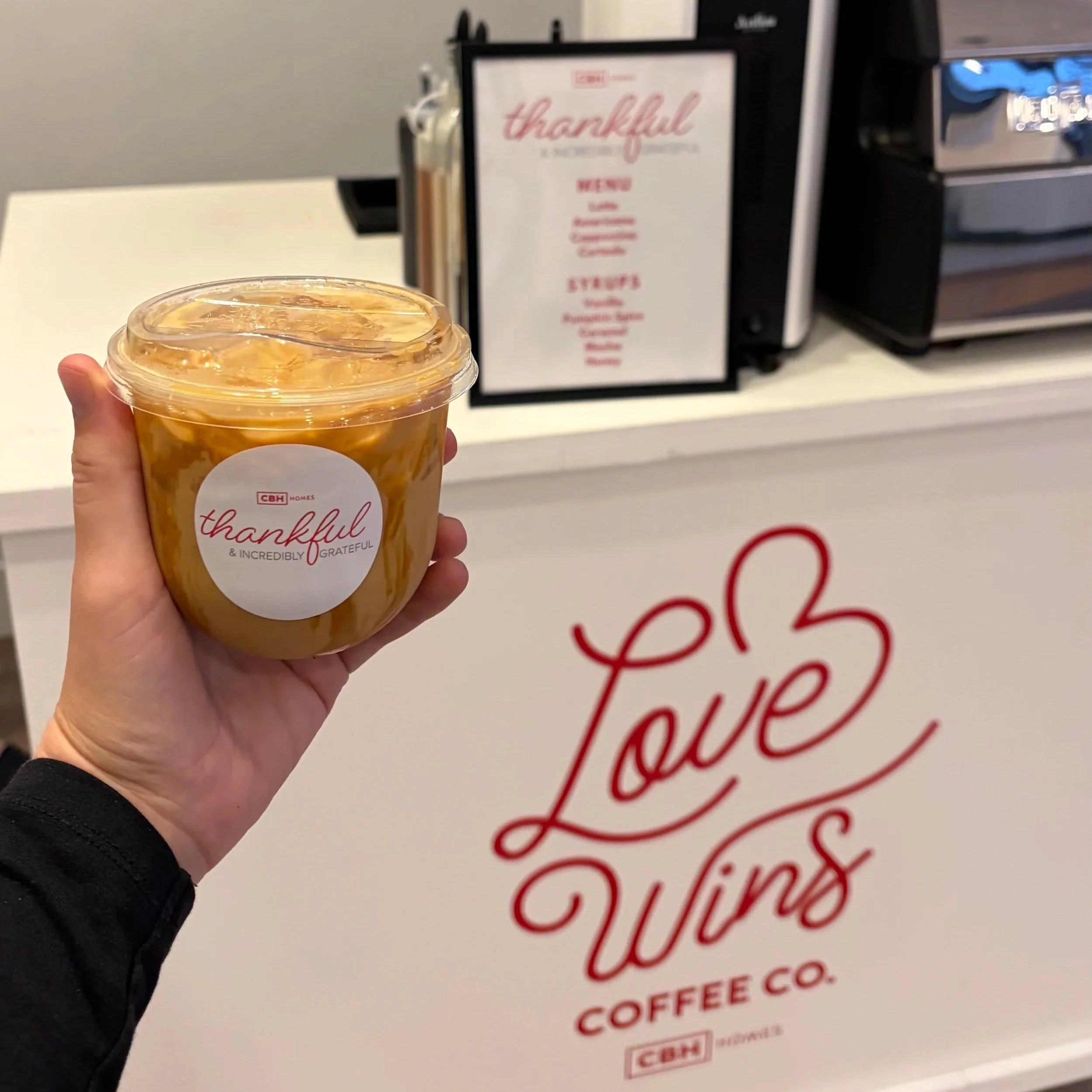A hand holding a custom branded coffee cup of iced coffee with a lid, branded with a 'thankful & INCREDIBLY GRATEFUL' sticker, in front of a white mobile coffee catering cart