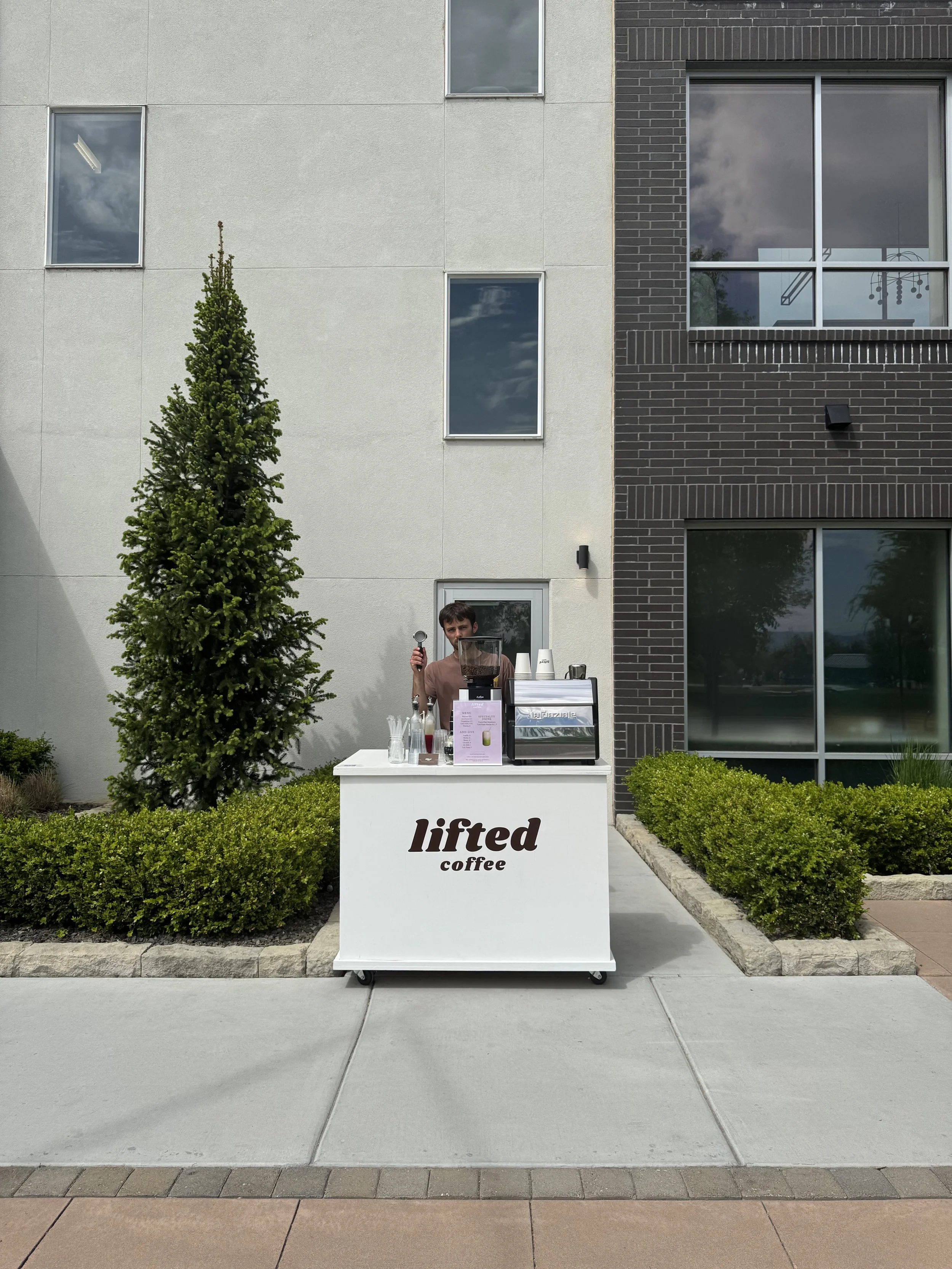 A person standing behind a small white coffee cart labeled lifted coffee. Mobile Specialty Coffee Cart and Espresso Bar Catering Boise Idaho and the Treasure Valley