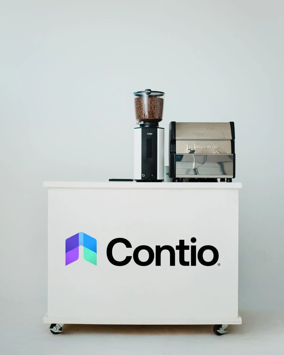 A coffee station with a coffee grinder filled with coffee beans, a commercial espresso machine, and a cart with a Contio logo on it. Custom corporate event for app and brand launch expo. 