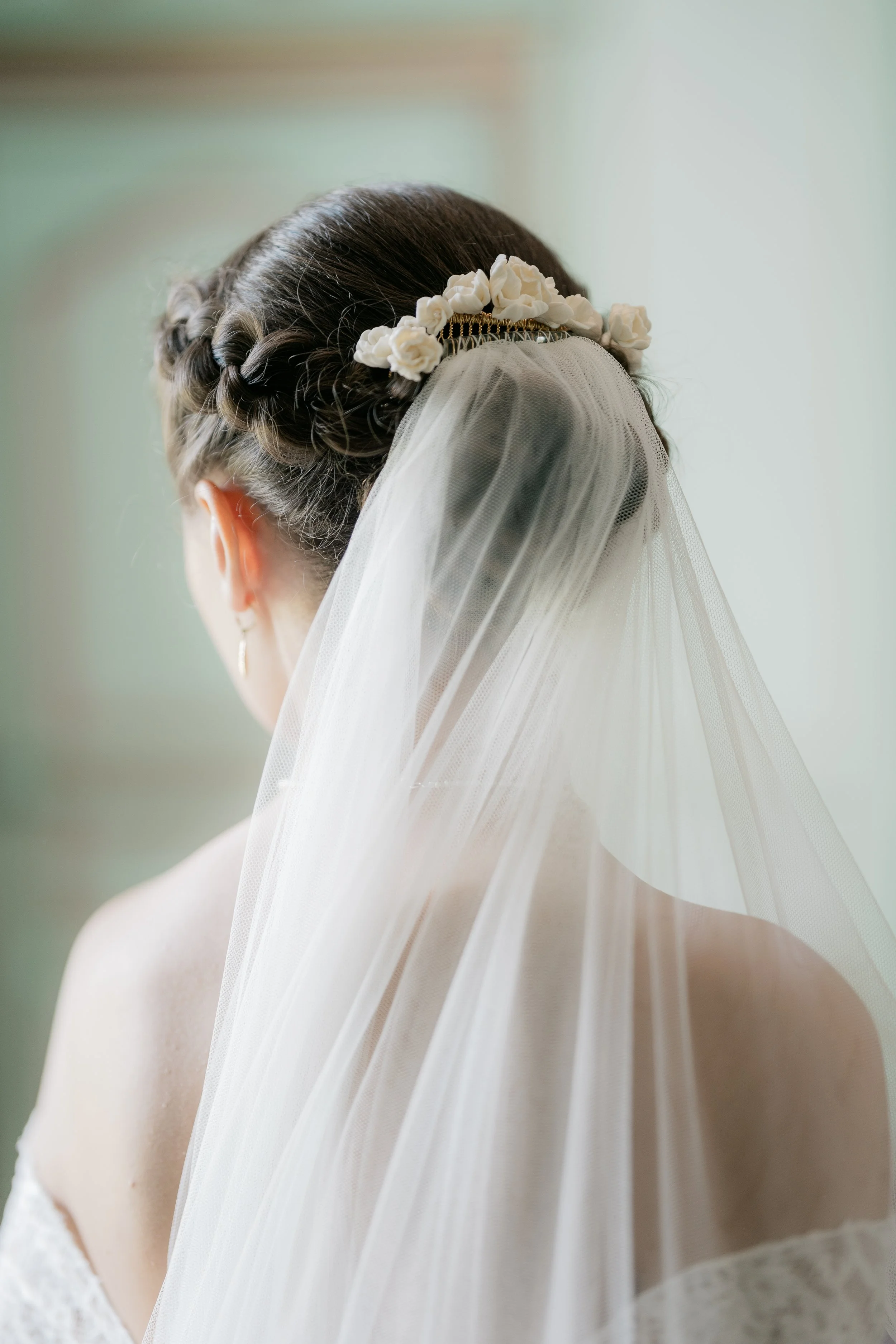 A bride with braided dark hair adorned with a white flower headband and a sheer white veil, viewed from behind, with a soft green background.