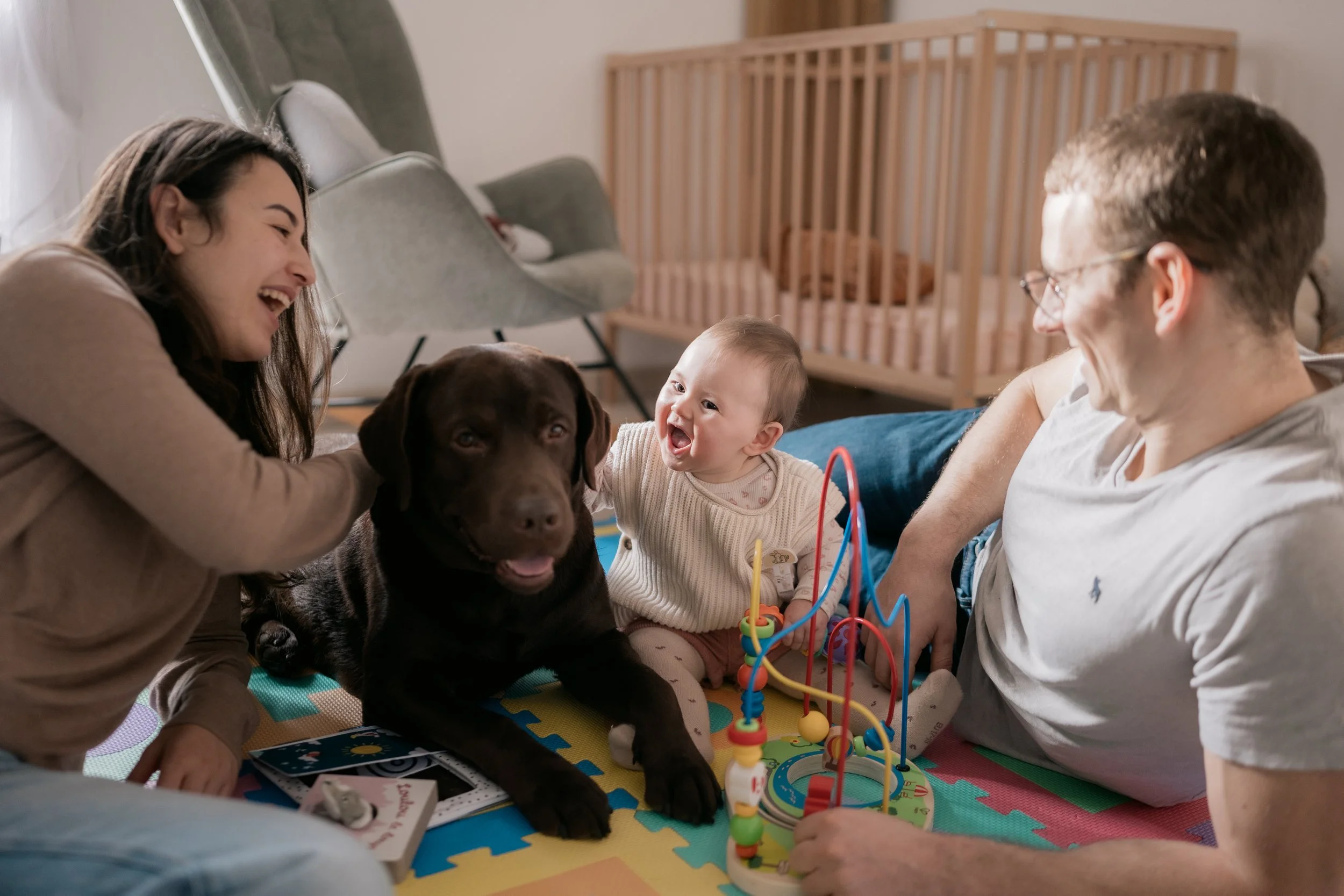A family with a woman, man, baby, and a Labrador retriever puppy playing together on a colorful foam play mat in a nursery with a wooden crib and modern chairs.
