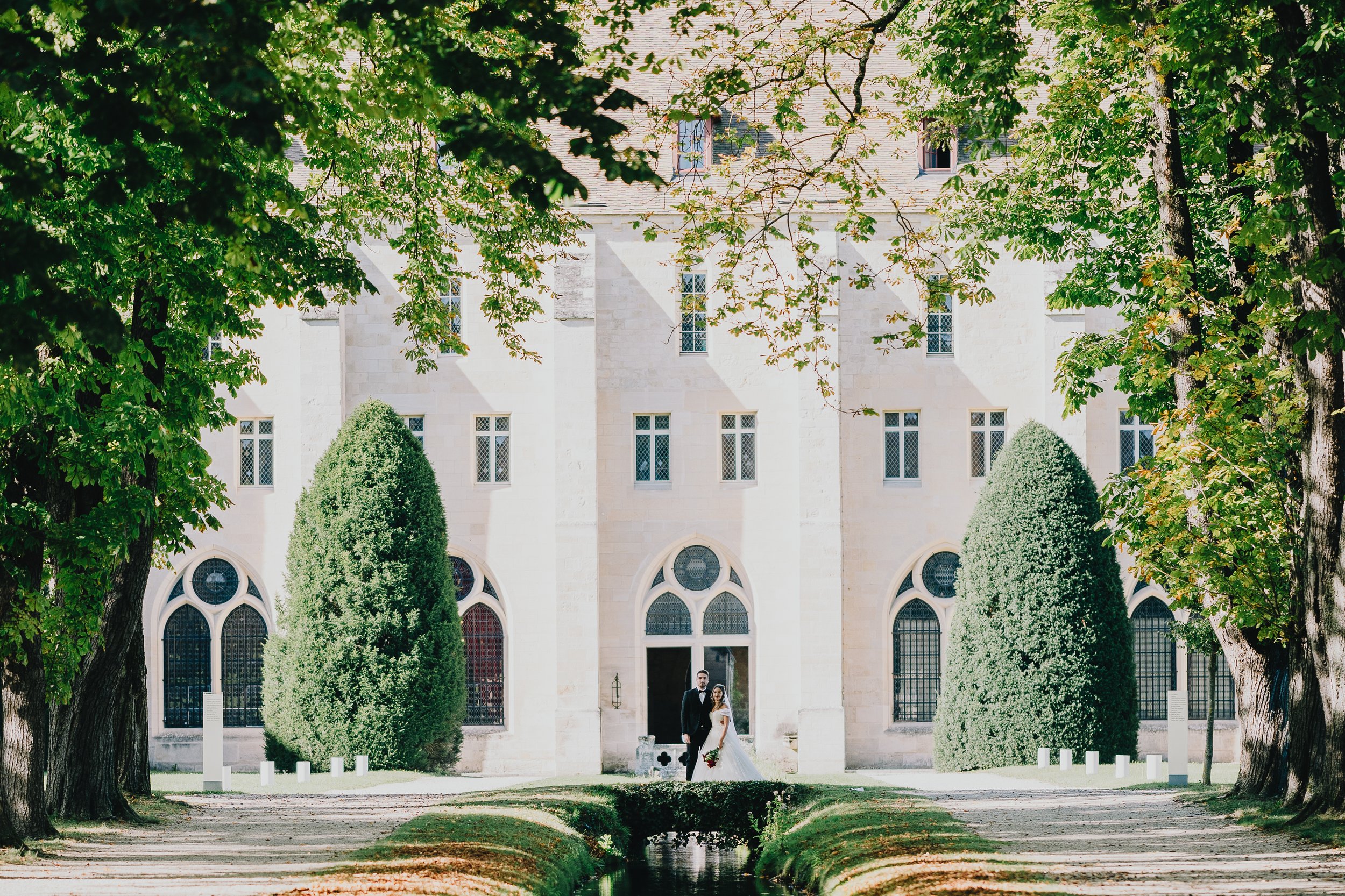 A bride and groom standing in front of a church, framed by large green trees and a small canal in the foreground.