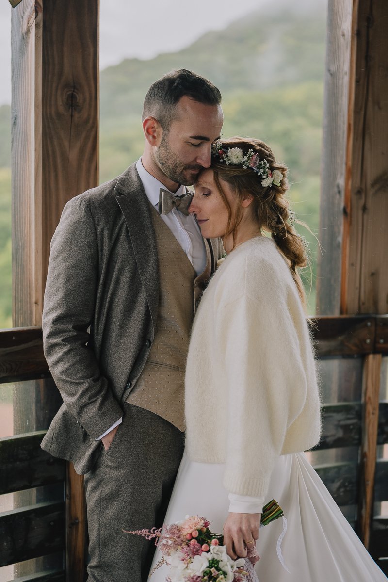 A bride and groom embrace on their wedding day, with the groom gently resting his forehead on the bride's head, inside a wooden structure with a view of a green landscape in the background.