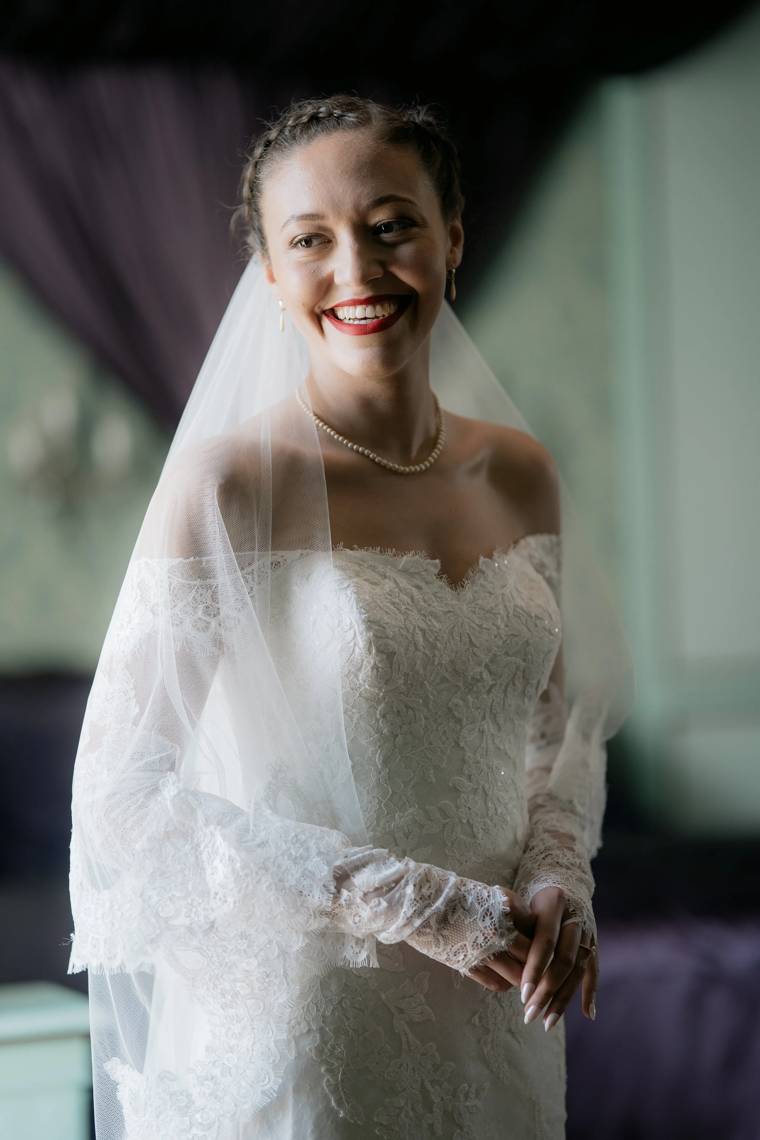 A smiling bride in a white lace wedding dress with a veil, pearl necklace, and earrings, standing indoors with soft natural light.