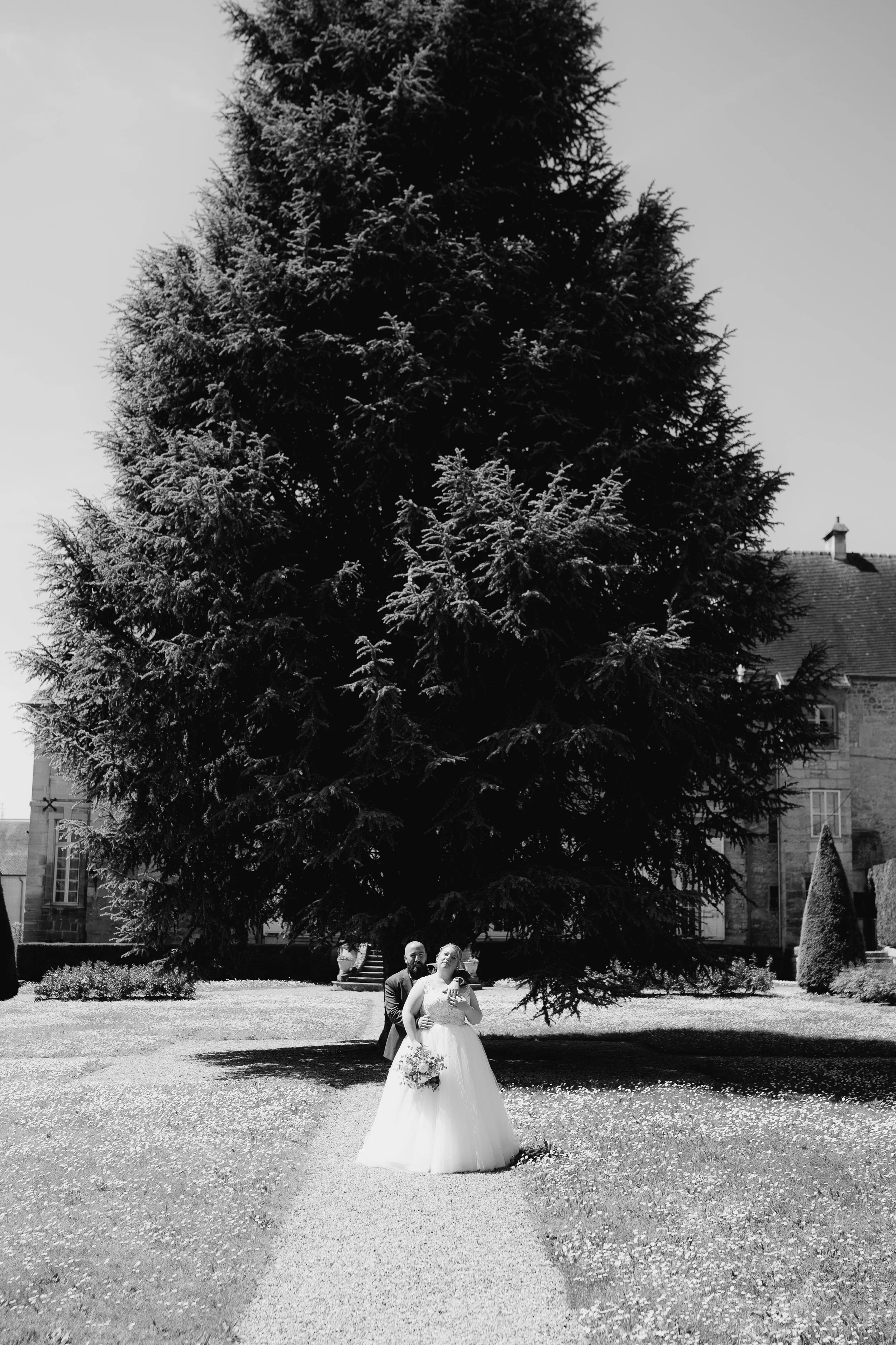 A black-and-white photo of a bride and groom standing in front of a large evergreen tree, with a building in the background, on a sunny day.