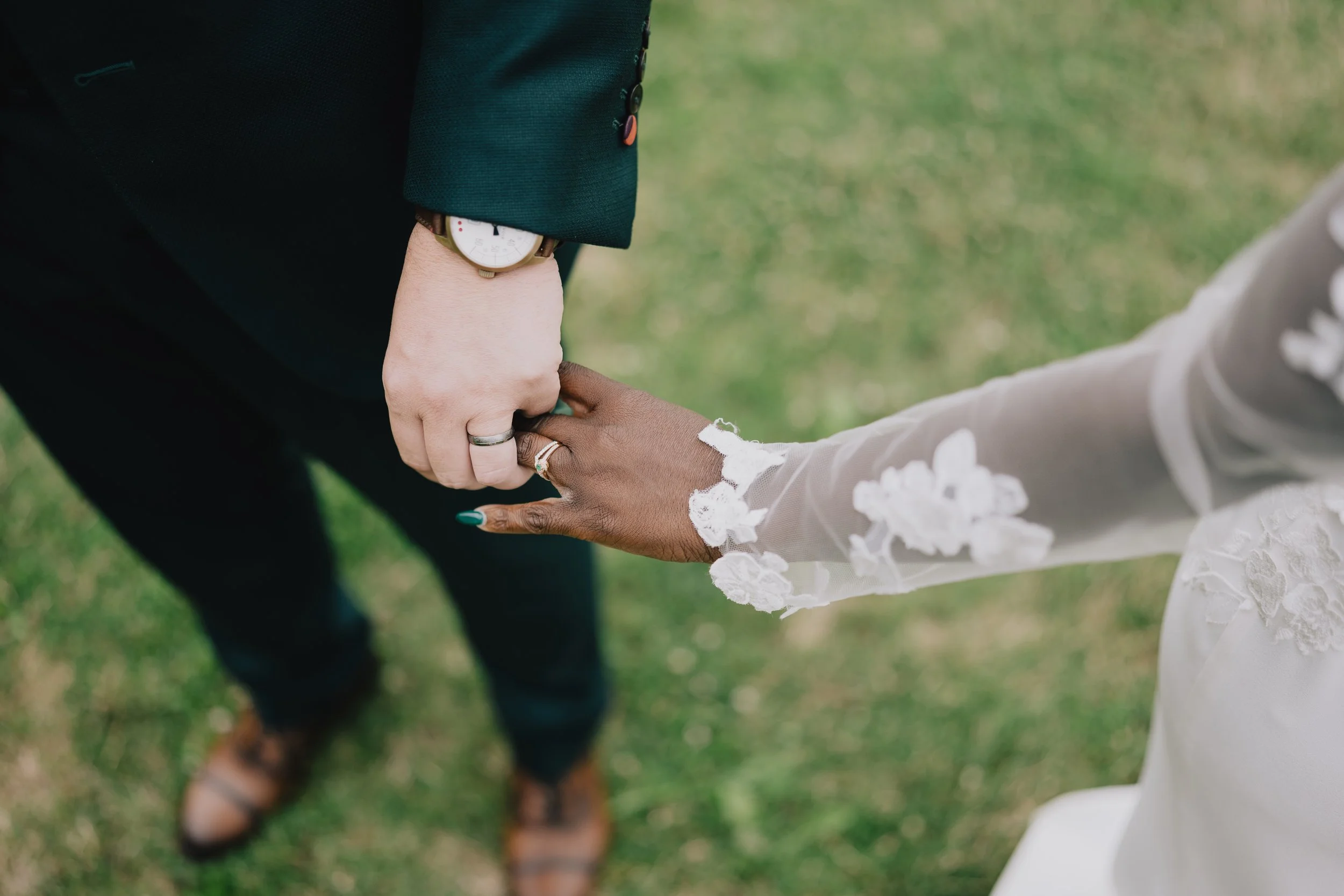 Close-up of a couple holding hands, with the woman wearing a white lace dress and silver rings, and the man dressed in a dark suit with a watch, standing on grass.