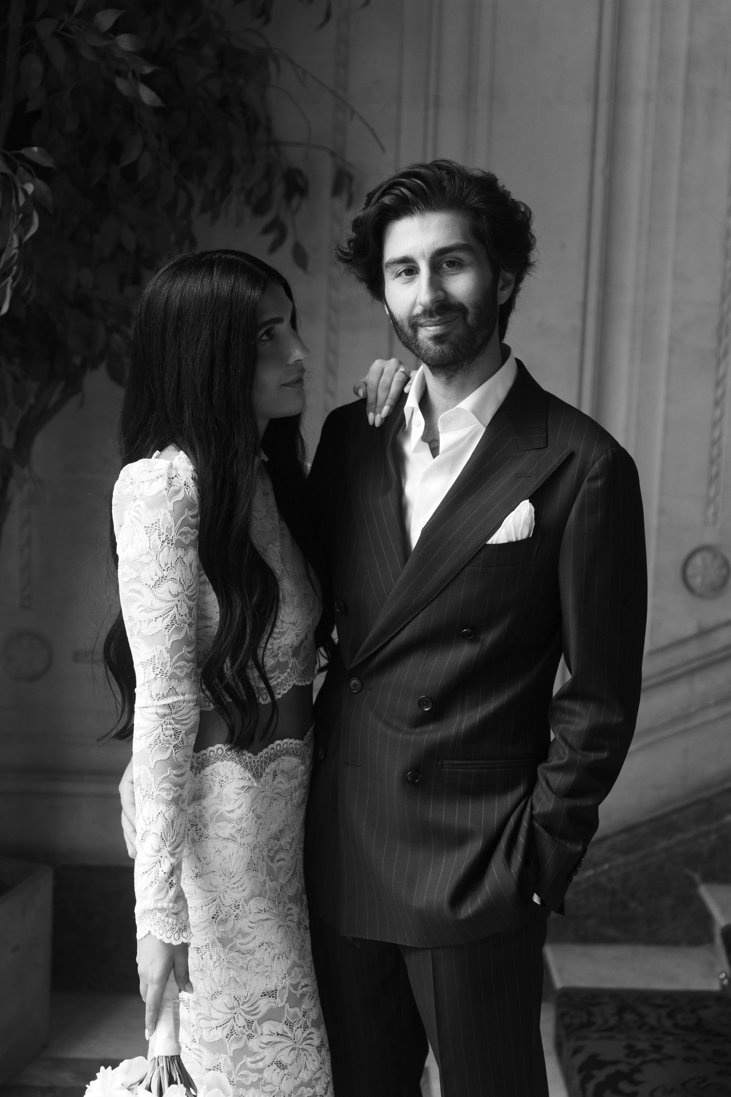 A black and white photo of a man and woman dressed in formal wedding attire. The woman wears a lace wedding dress and is holding a bouquet of flowers, while the man wears a pinstripe suit with a pocket square, and has a beard and wavy hair. They stan