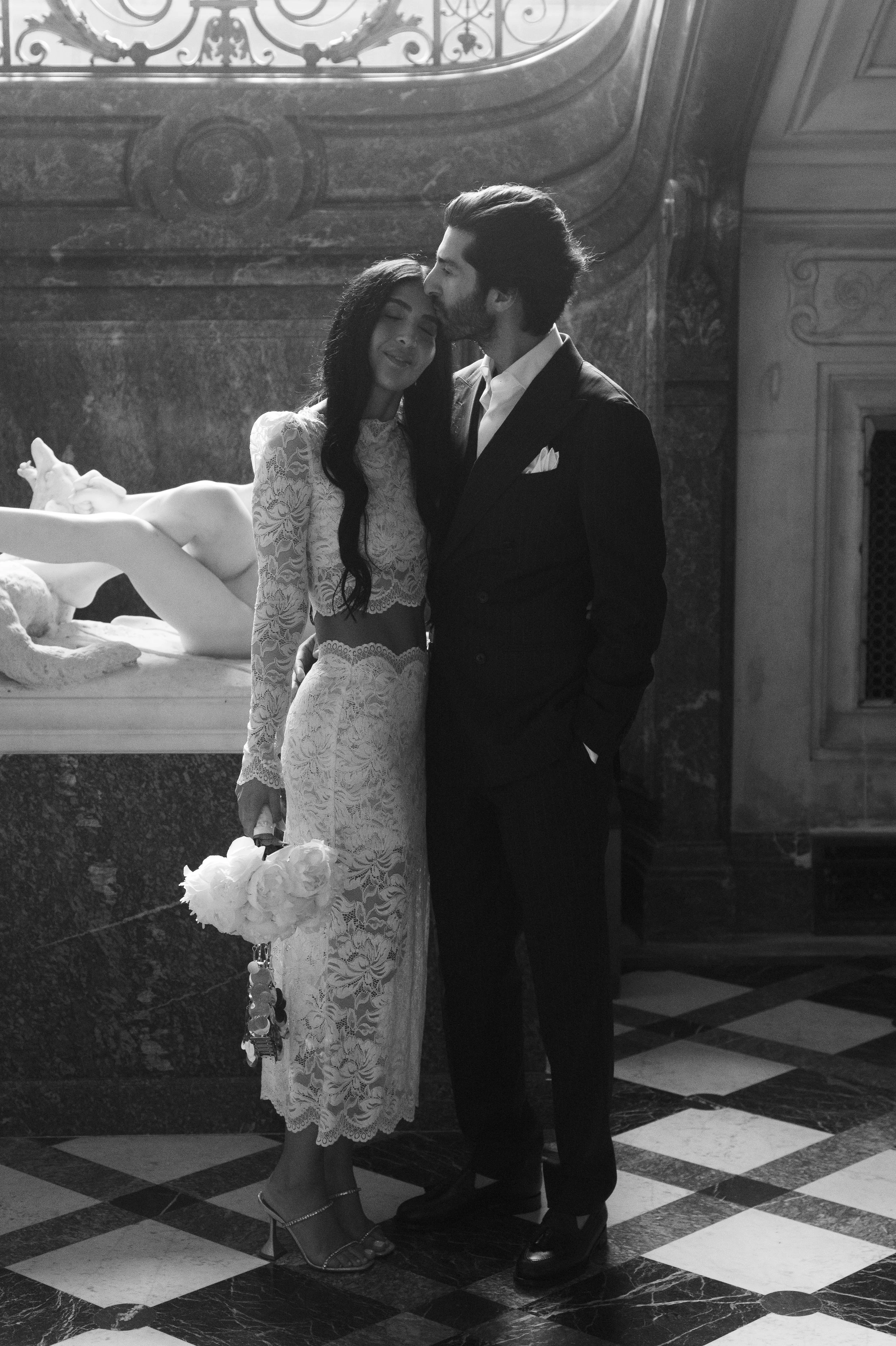 A black and white photo of a couple at a formal event, with the woman in a lace dress holding a bouquet of flowers, and the man in a tuxedo, kissing her on the forehead.