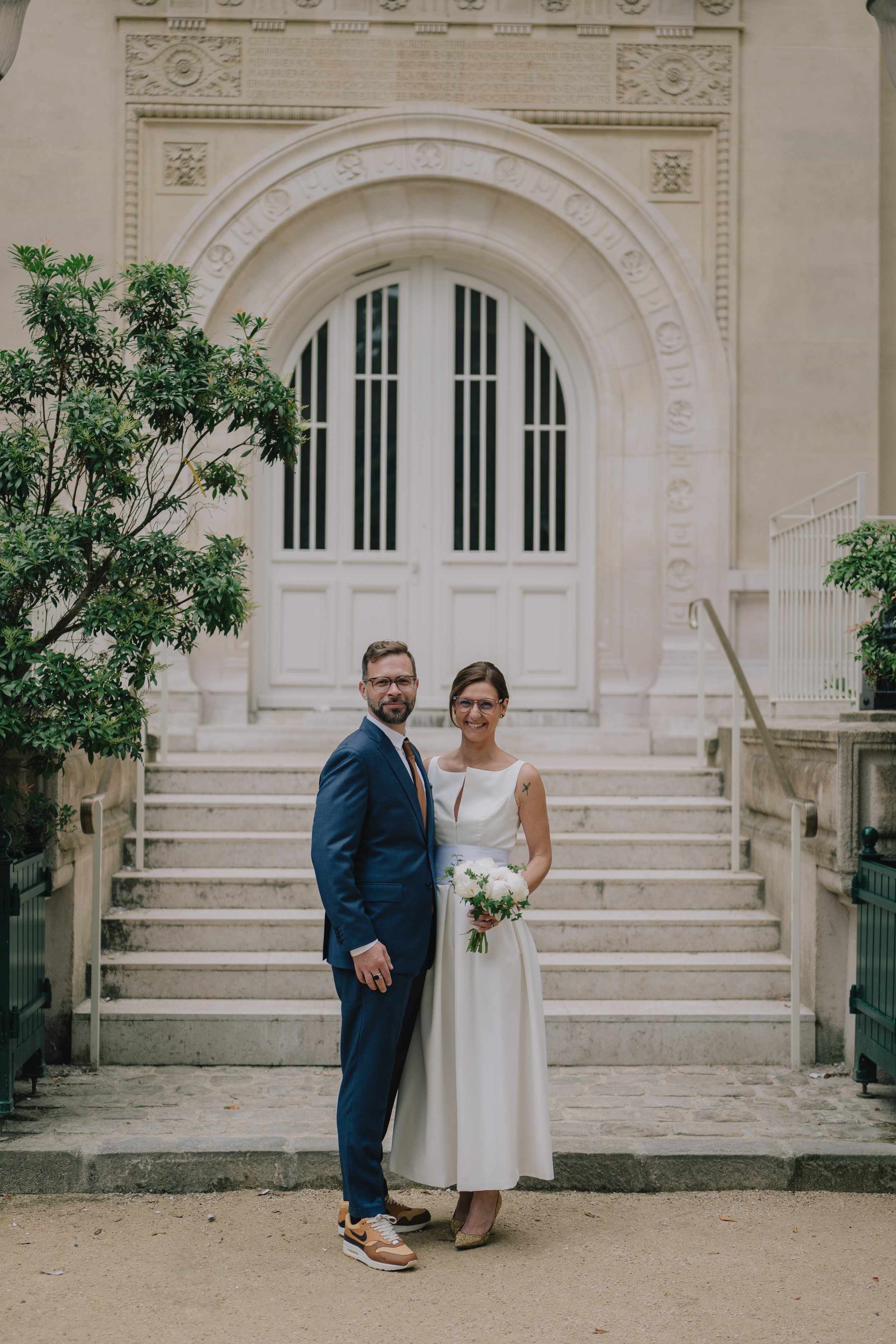 A newlywed couple standing on steps in front of a historic building with an arched doorway, greenery on the sides, and stone steps, smiling and posing for a photo.