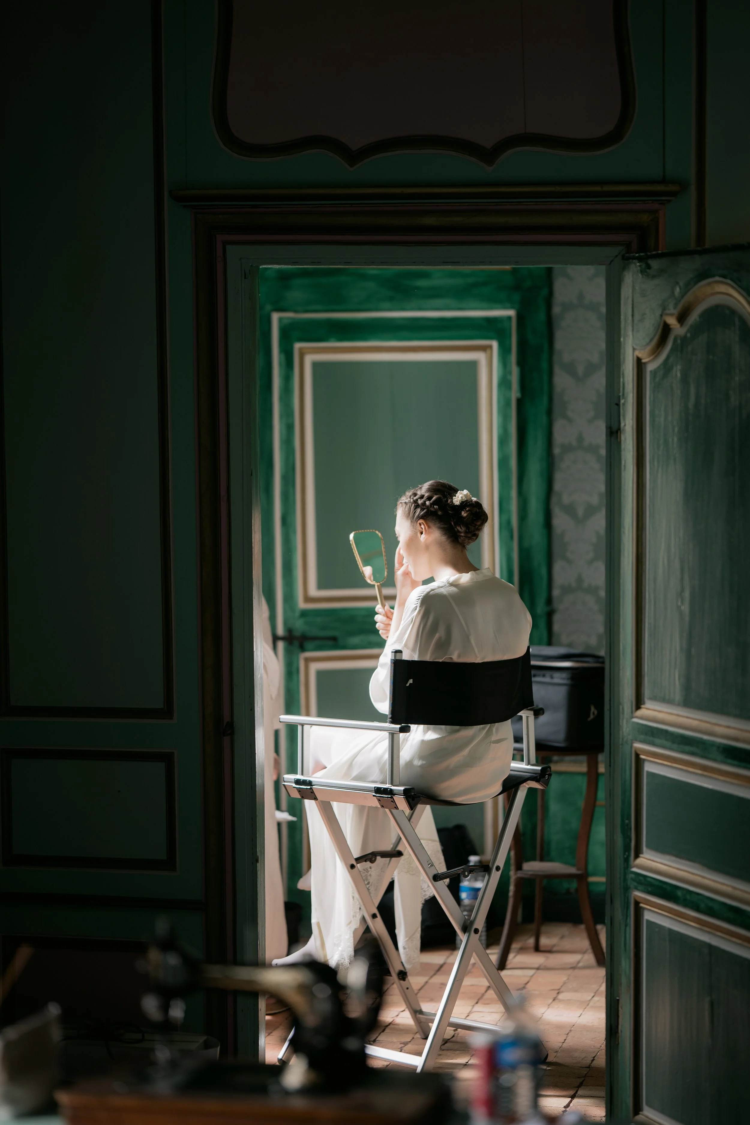 Woman in white sitting on makeup chair looking into mirror in a green-walled room.