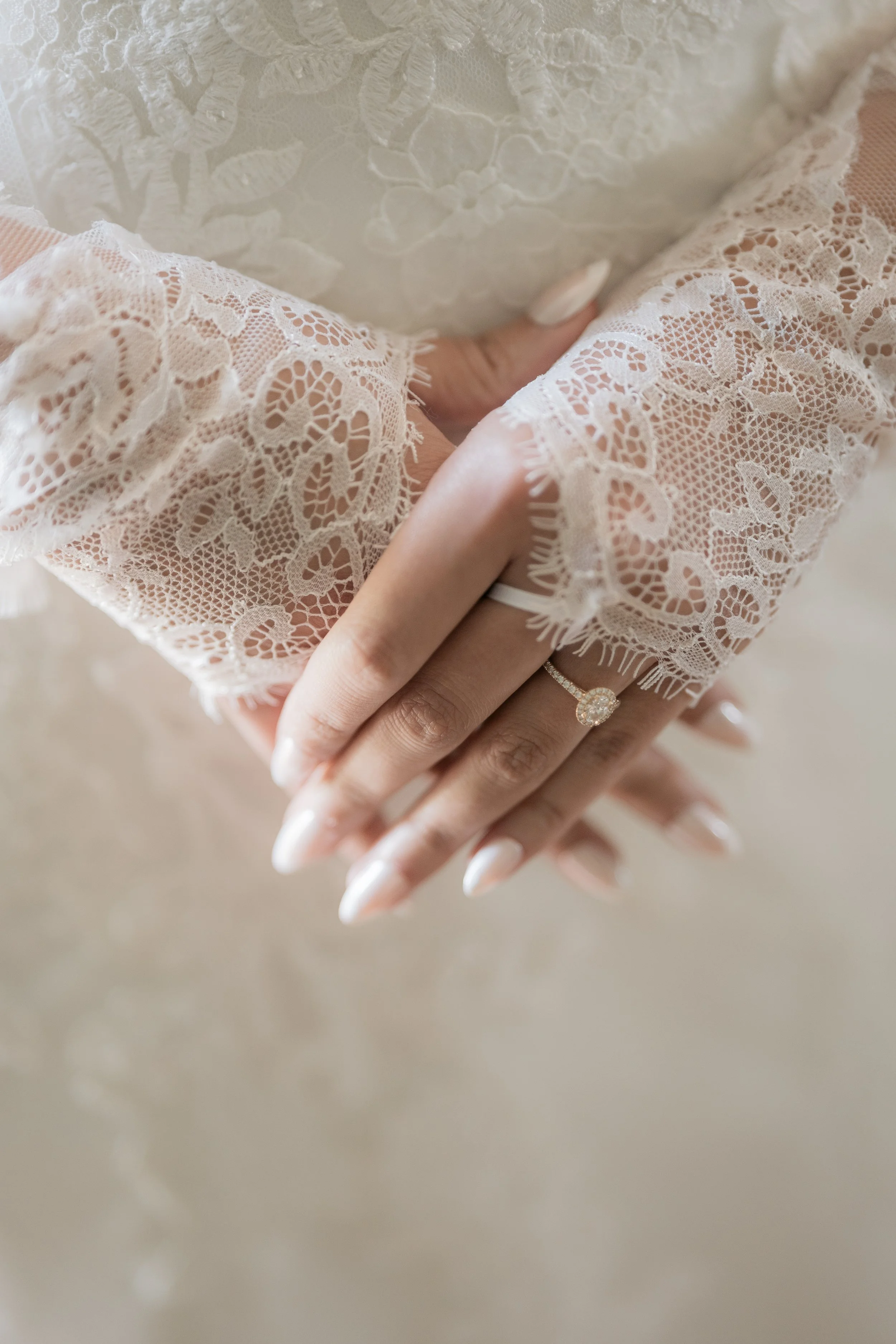 Close-up of two hands with elaborately decorated lace sleeves, one hand wearing a diamond engagement ring.