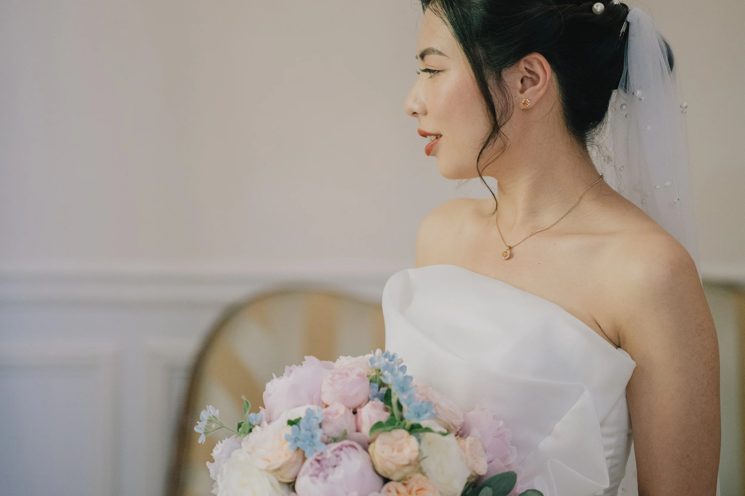 A bride in a strapless white dress holding a bouquet of pastel pink, white, and light blue flowers, with a veil attached to her dark hair styled in an elegant updo, smiling with eyes closed.