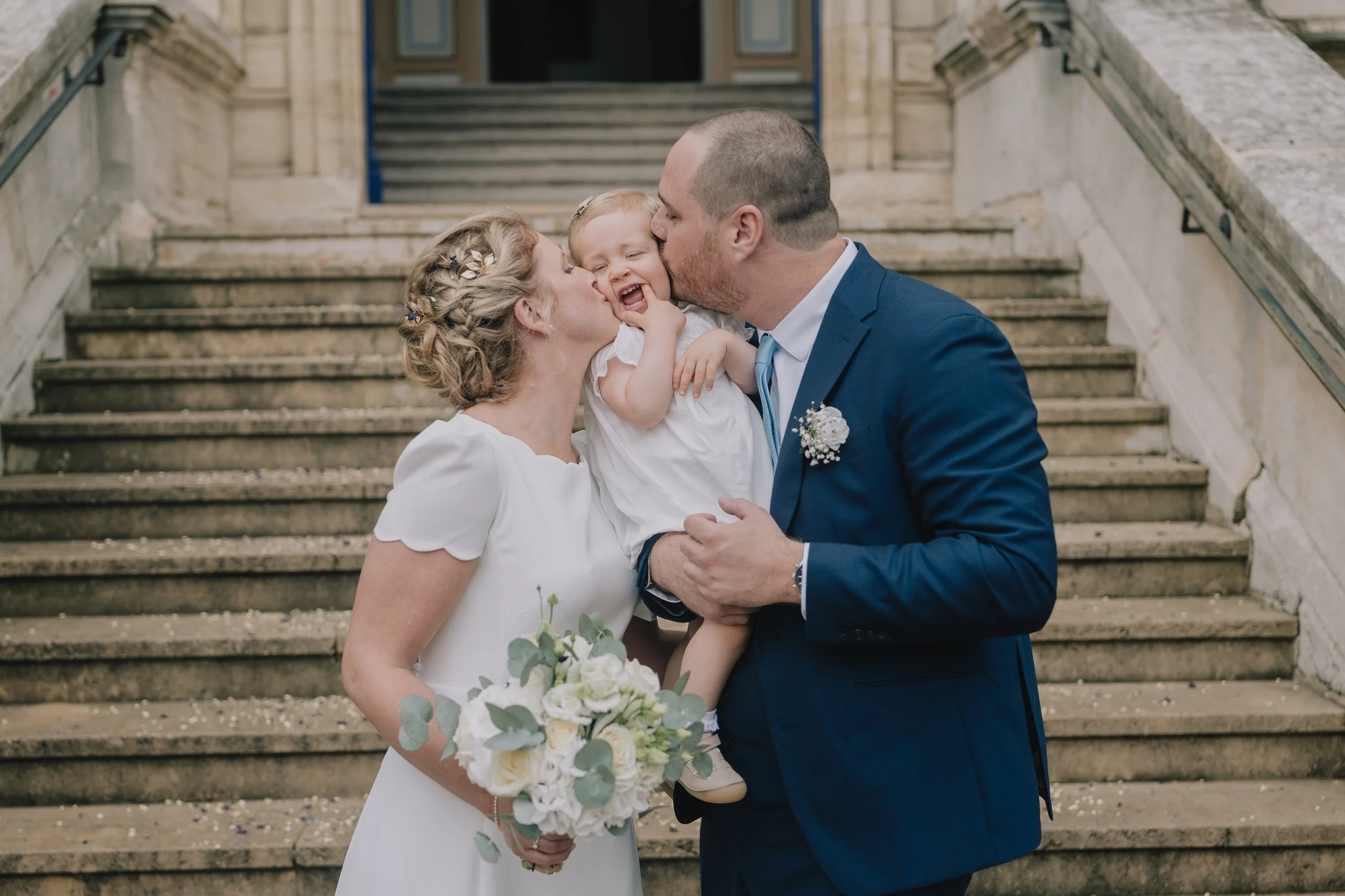 A bride and groom holding a young child and sharing a kiss on the steps outside a building, during a wedding celebration.