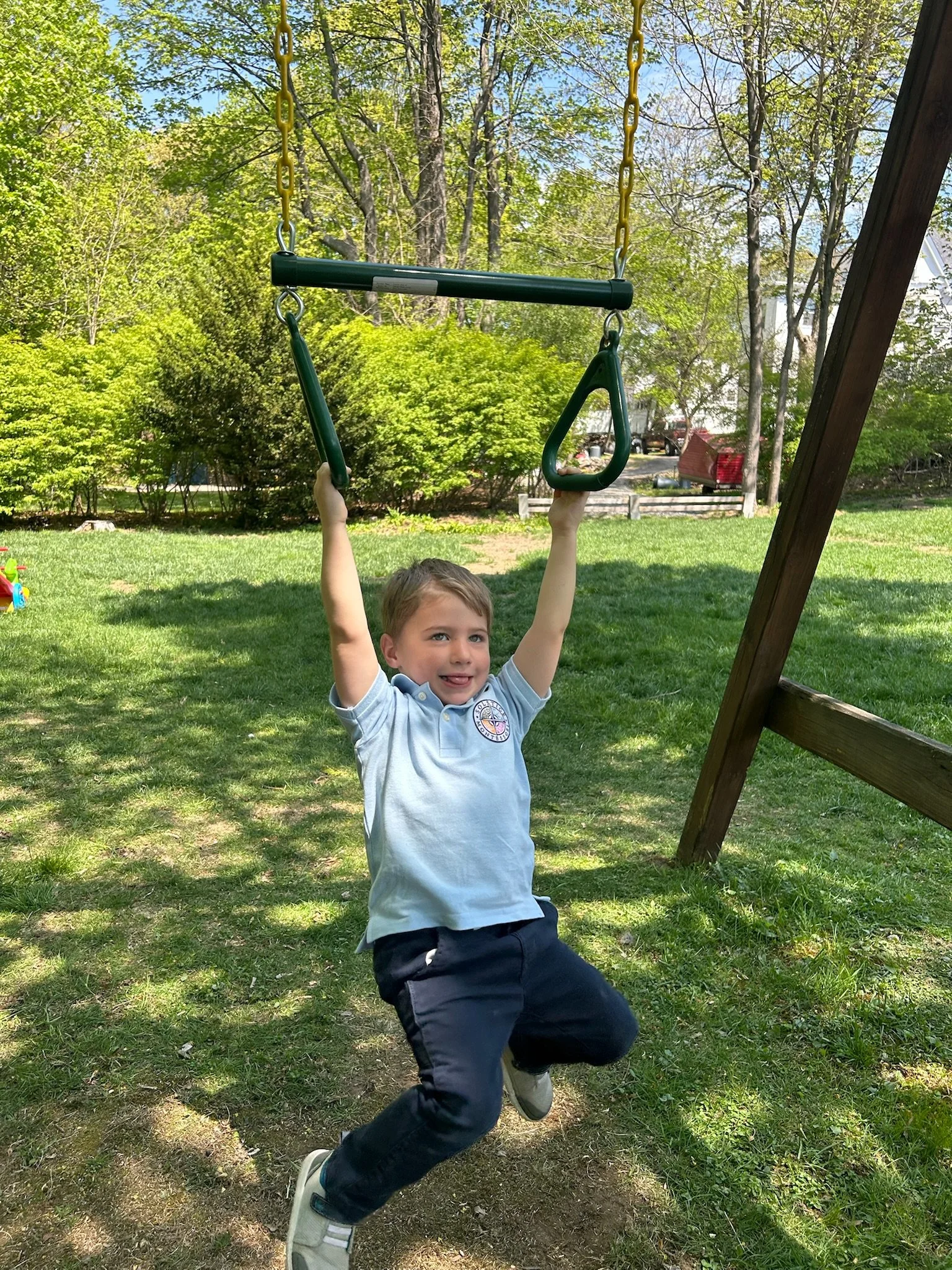 Child holding onto a swing set with both hands, mid-air, in a grassy backyard area with trees and a clear sky.