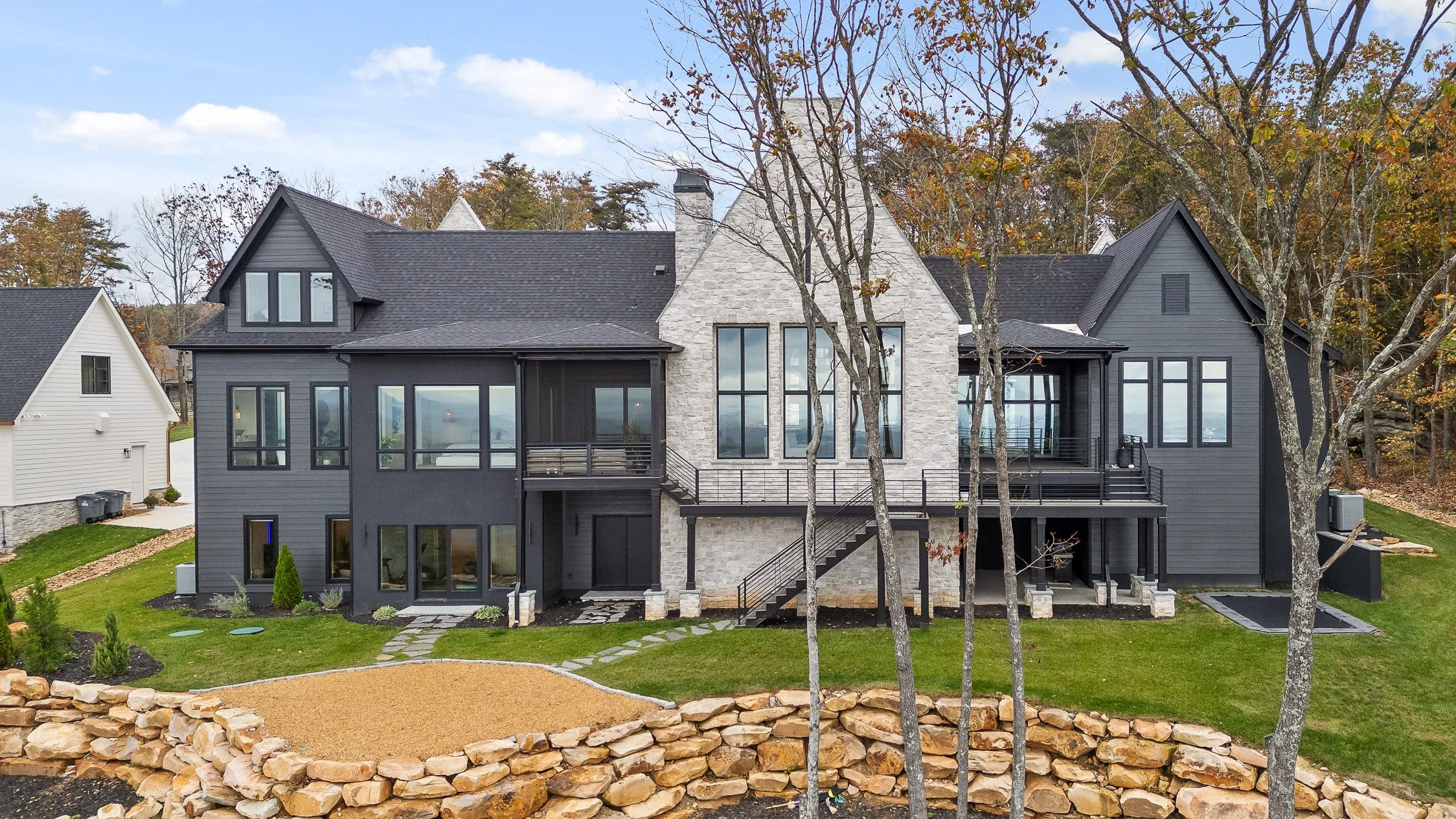 Large modern house with dark gray and white stone exterior, multiple large windows, and multiple decks, set on a landscaped yard with trees and a stone retaining wall.
