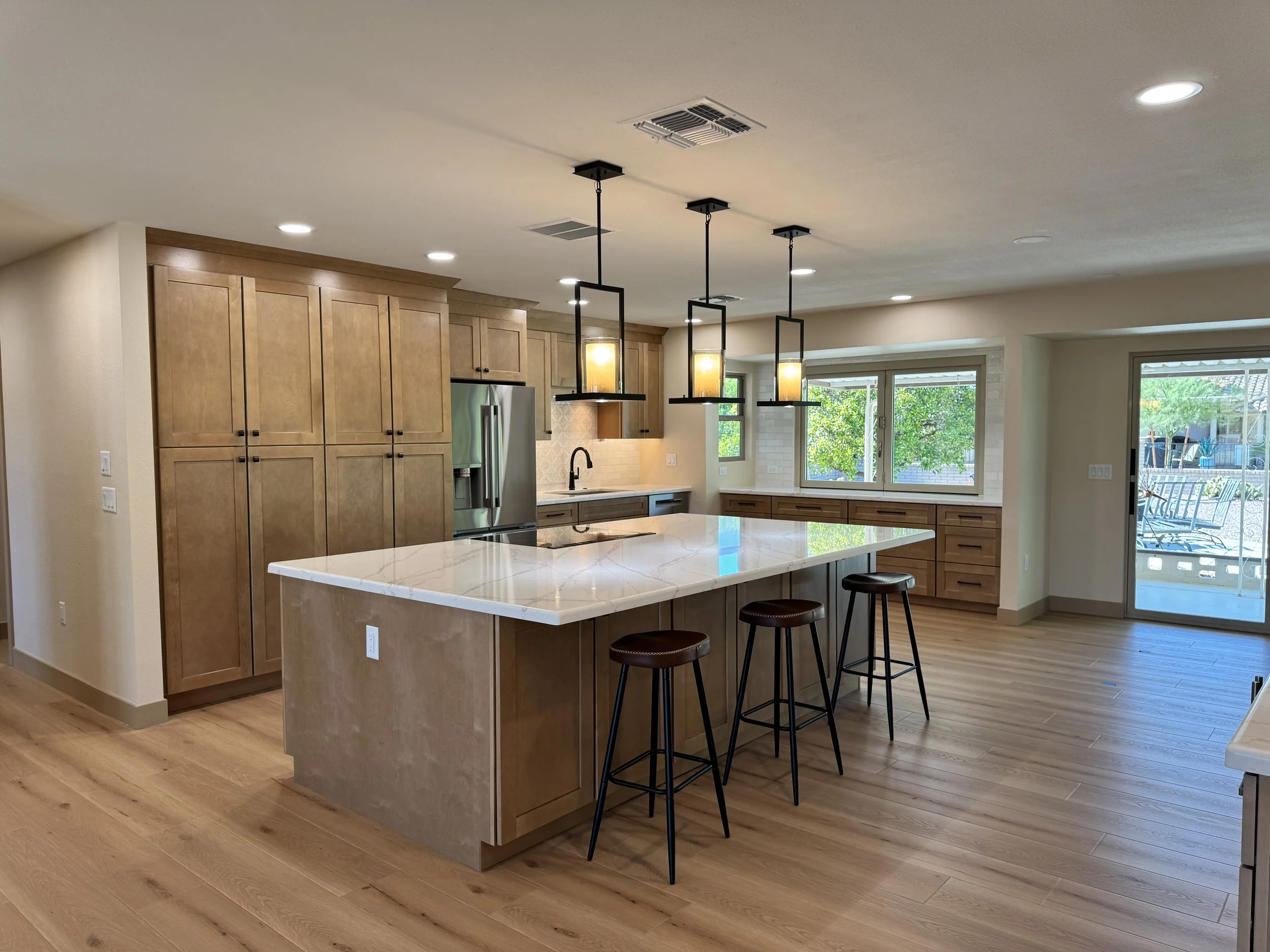 Modern kitchen with wooden cabinets, a large white marble island, three black bar stools, pendant lights, and large windows showing outdoor greenery.