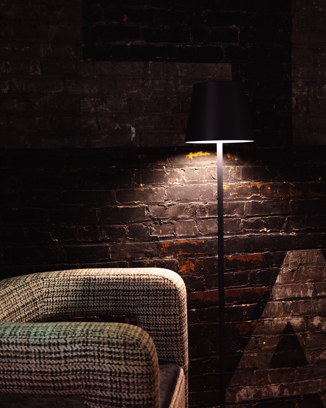 A black floor lamp with a cone-shaped shade illuminating a textured fabric armchair in front of a dark brick wall.