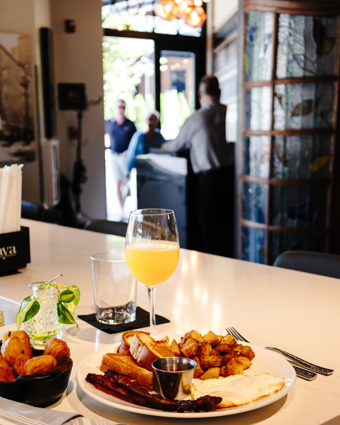 A breakfast plate with eggs, bacon, toast, and roasted potatoes on a white table, with a glass of orange juice and a glass of water. In the background, a restaurant or cafe interior with people and a server near the entrance.