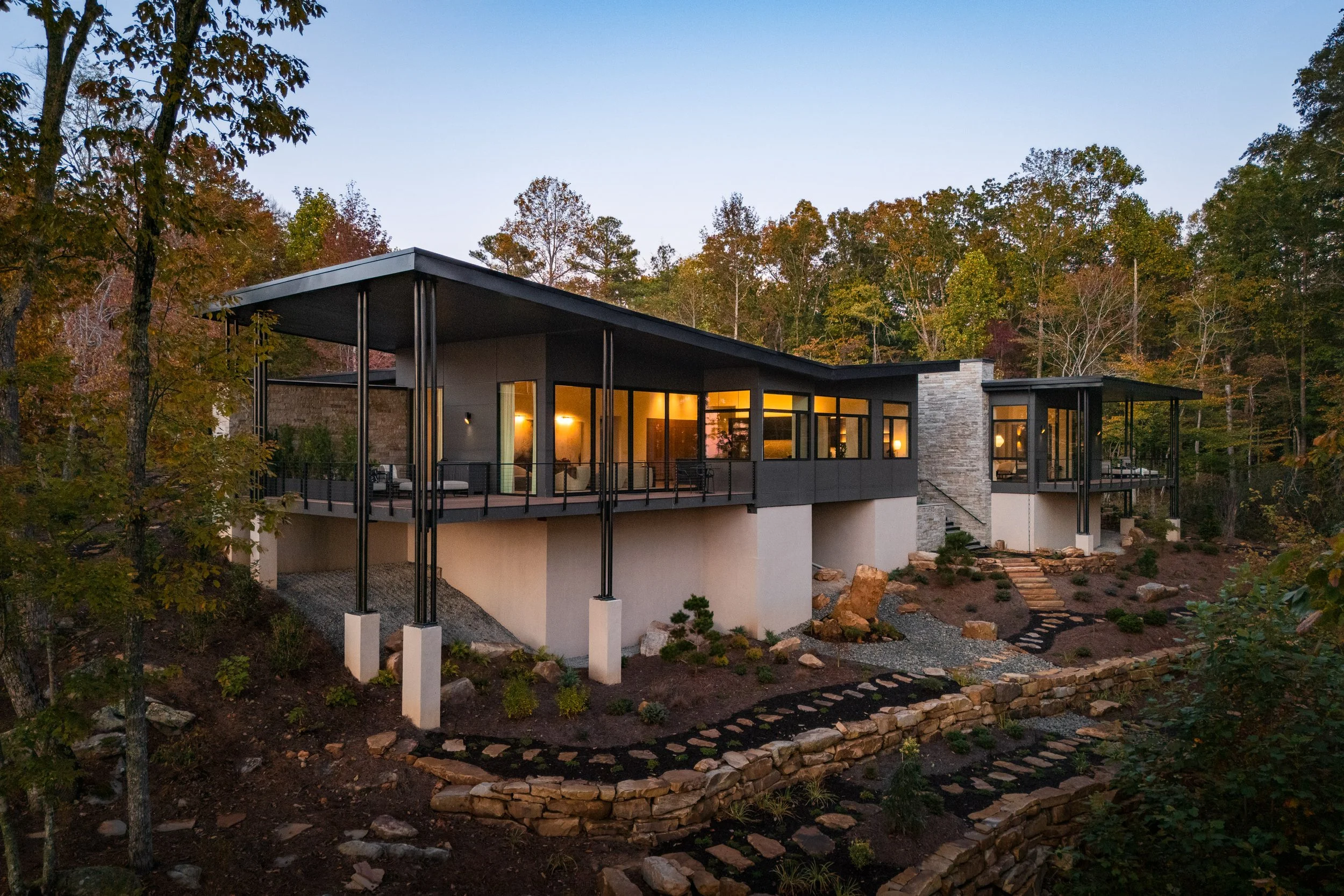 Modern house on a hillside at dusk, with large windows and a spacious balcony, surrounded by trees and landscaping.