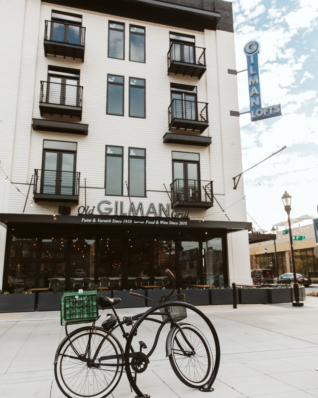 A black bicycle with a green crate attached to the back parked on a sidewalk in front of a modern building with balconies and signage for Old Gilman Grill and GILMAN LOFTS.