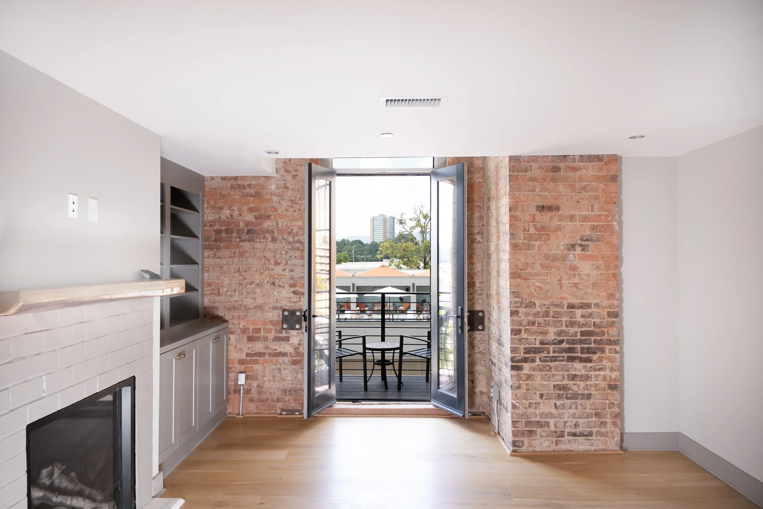 Interior of a room with brick walls and hardwood floor, open double glass doors leading to a balcony with a small table and chairs, cityscape visible in the background.
