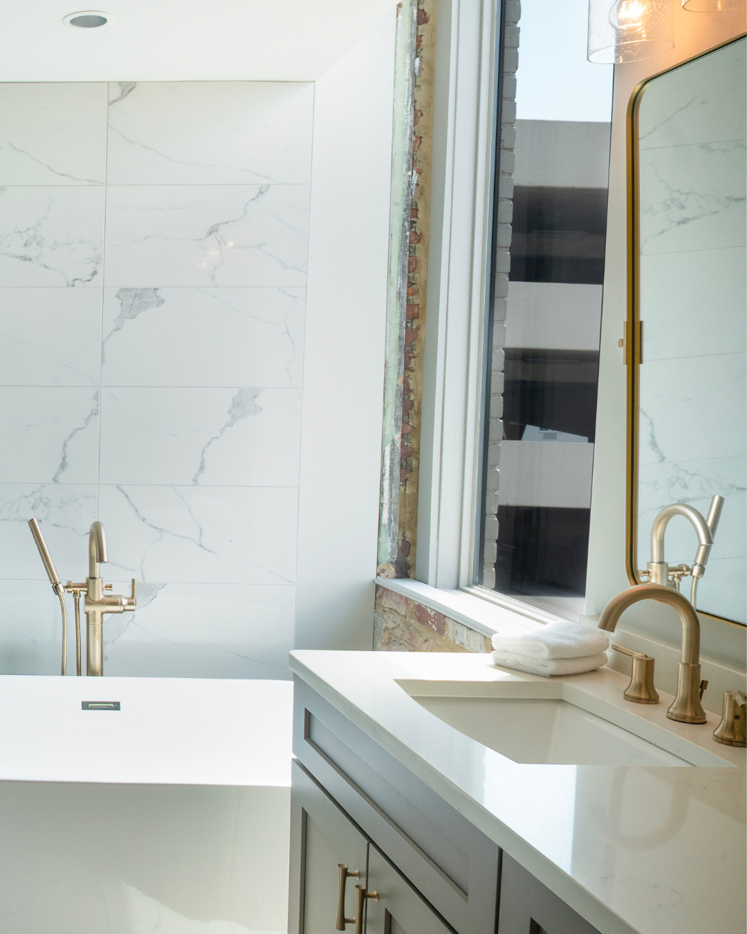 Modern bathroom with white marble wall tiles, a white bathtub, a brass faucet, a white countertop with folded towels, and a window showing an adjacent brick wall.