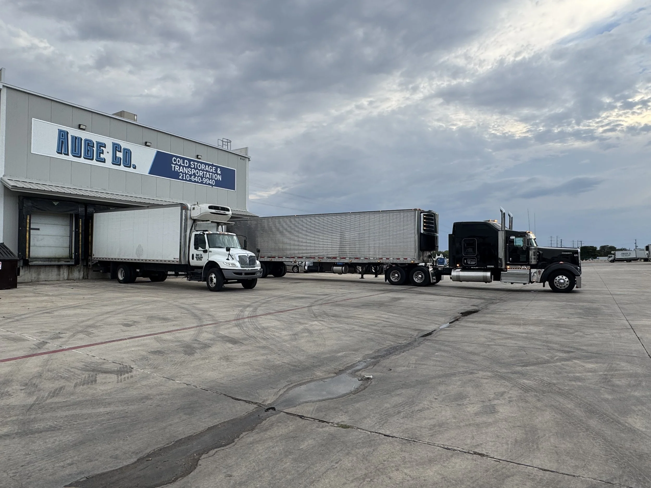 A large industrial building with a blue sign reading 'AUGE CO. Cold Storage & Transportation' and a phone number. Several semi-trucks are parked outside on a concrete lot, with overcast sky in the background.