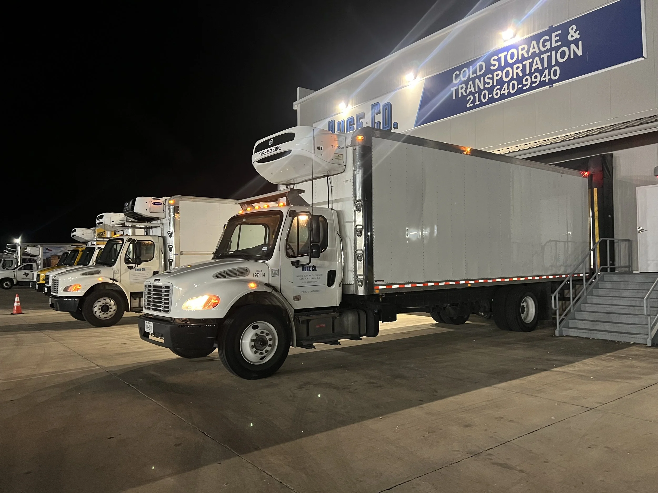 Nighttime view of a row of refrigerated trucks parked outside a storage and transportation facility, with a sign indicating cold storage services.
