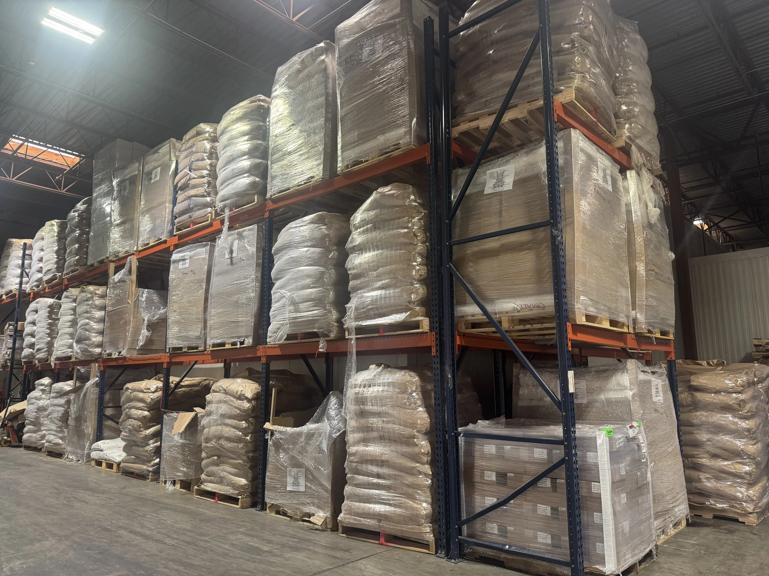 Stacked pallets of boxed and wrapped goods stored on industrial metal shelving inside a warehouse.