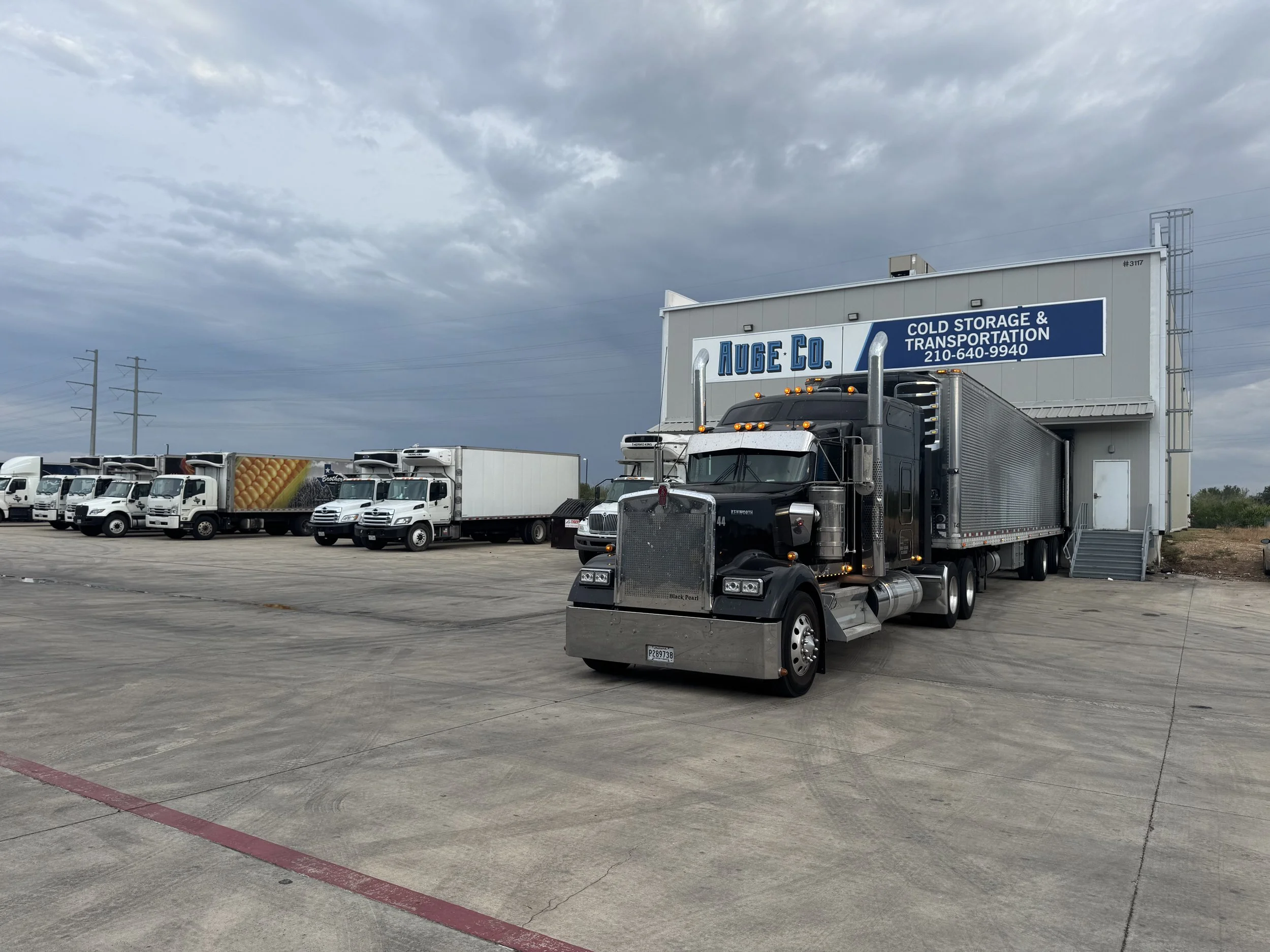 A large black semi-truck parked outside a warehouse with a sign reading 'Auge Co. Cold Storage & Transportation'. Several other white trucks are lined up in the background under a cloudy sky.