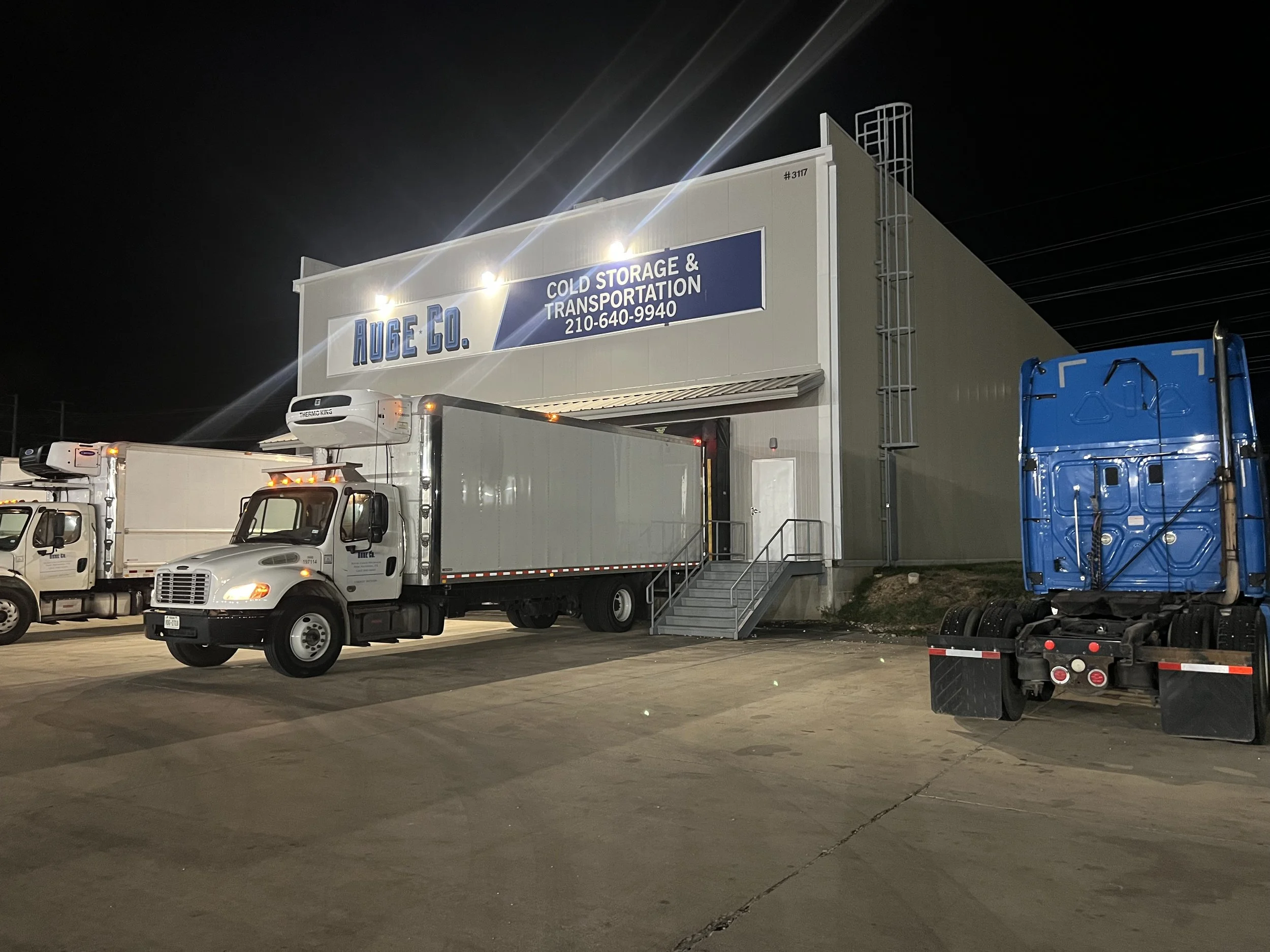 Large warehouse building with a sign reading 'AUGE CO. Cold Storage & Transportation,' daytime trucks parked outside at night.