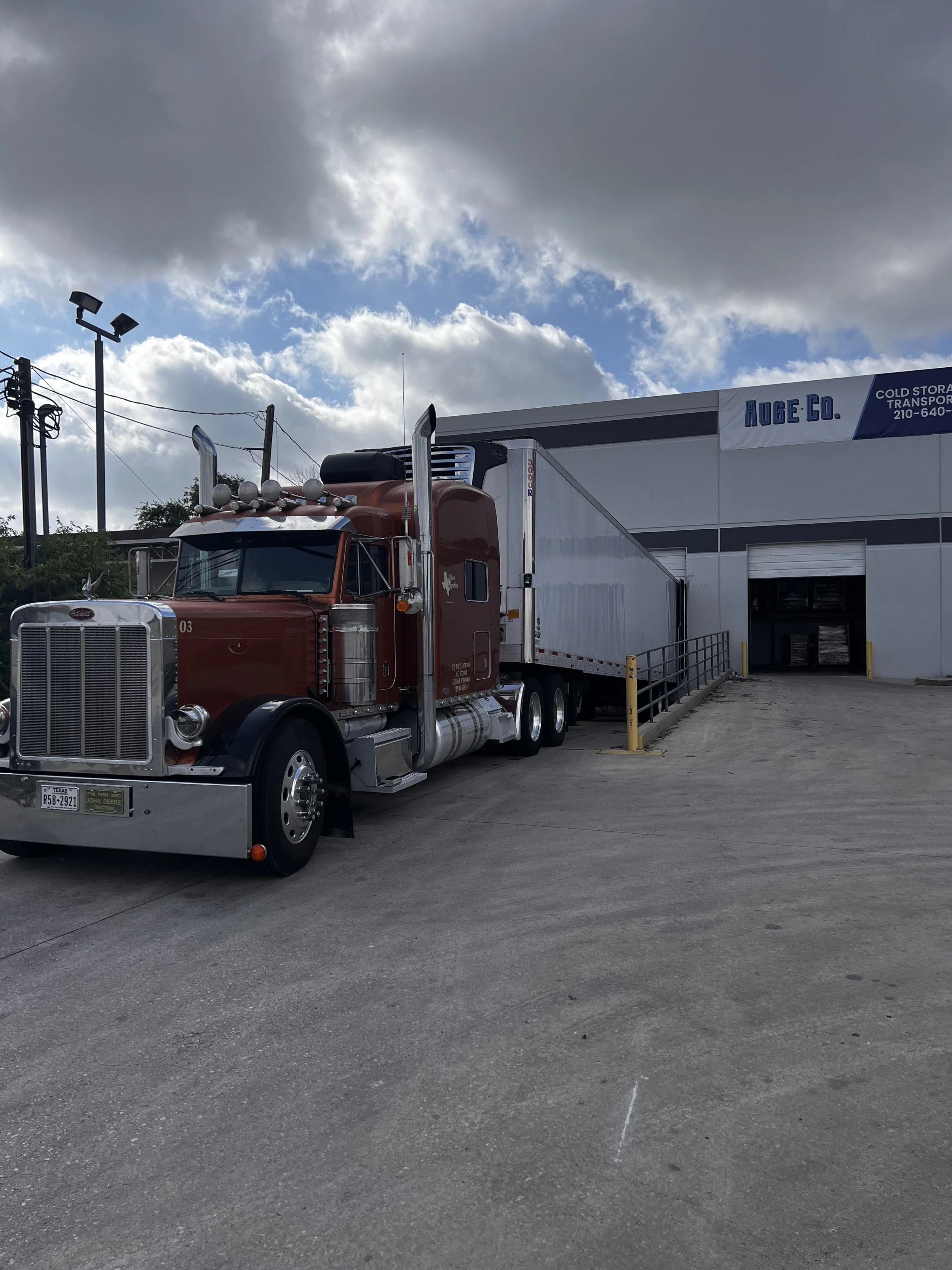 A semi-truck with a red cab parked next to a loading dock at a warehouse with a sign that reads 'Auge Co.' and a cloudy sky above.