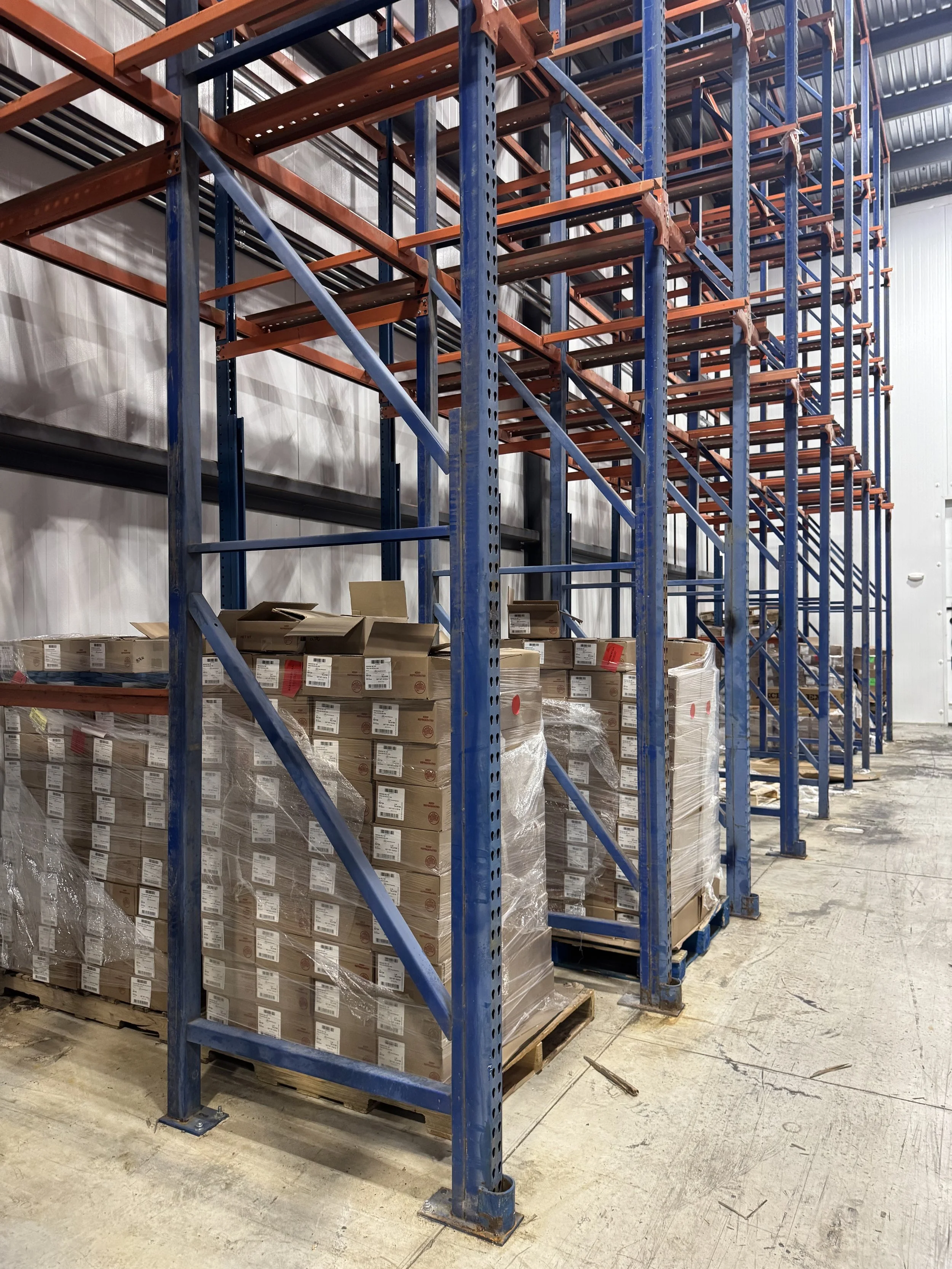 Empty industrial warehouse with blue and orange metal shelving racks, stacked with boxes wrapped in plastic on pallets, concrete floor, and white walls.