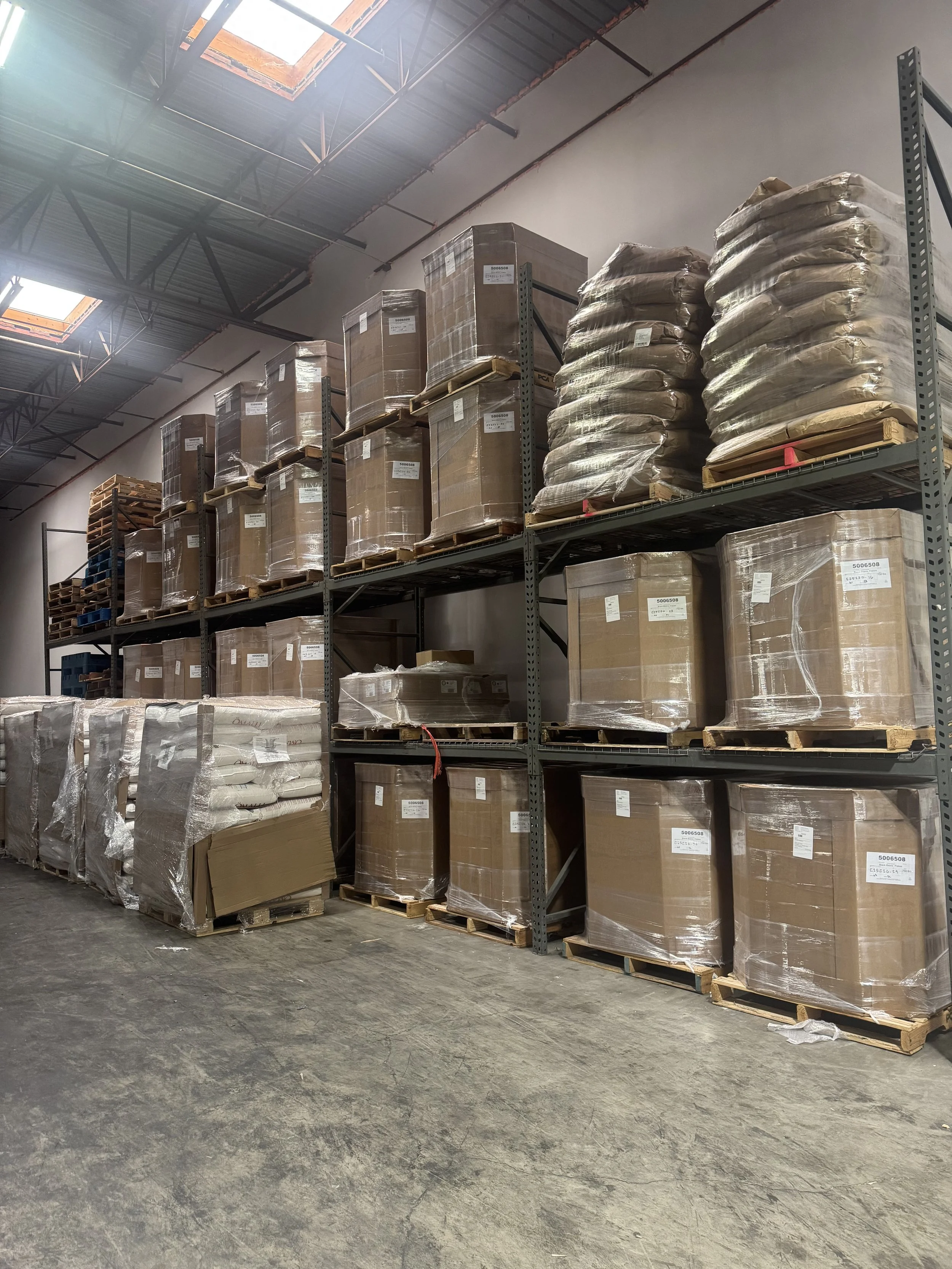 Shelves stocked with pallets of boxed and wrapped goods in a warehouse. Skylights in the ceiling provide natural light.