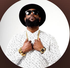 Man in hat and sunglasses wearing a white patterned shirt with gold accessories