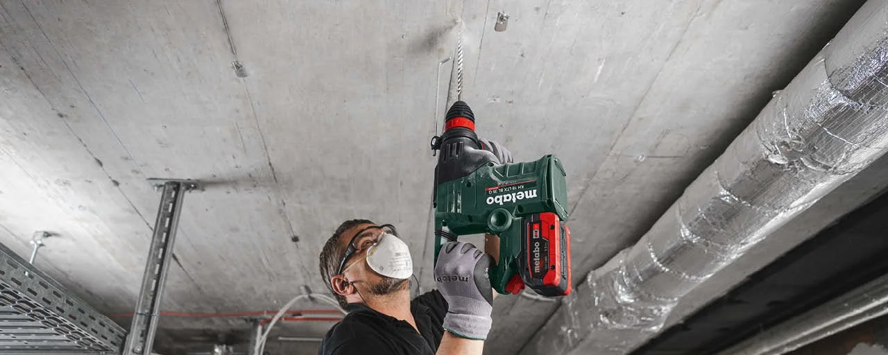 A construction worker wearing safety goggles and a face mask is using a cordless drill to work on the ceiling of a building under construction.
