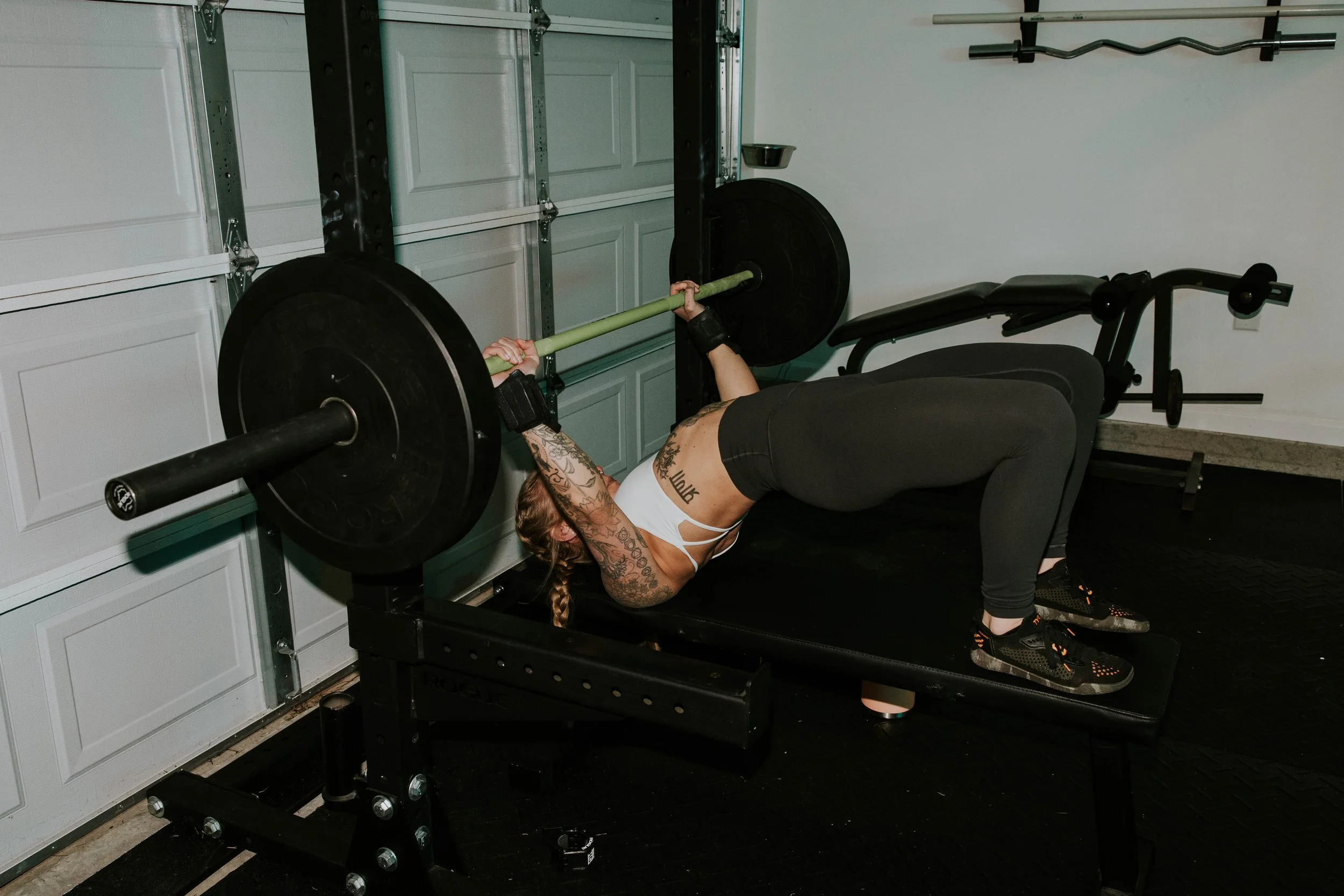 A woman with tattoos on her arm and a ponytail lying on a workout bench, performing a bench press with a barbell with black weight plates, in a garage gym with a white garage door in the background.