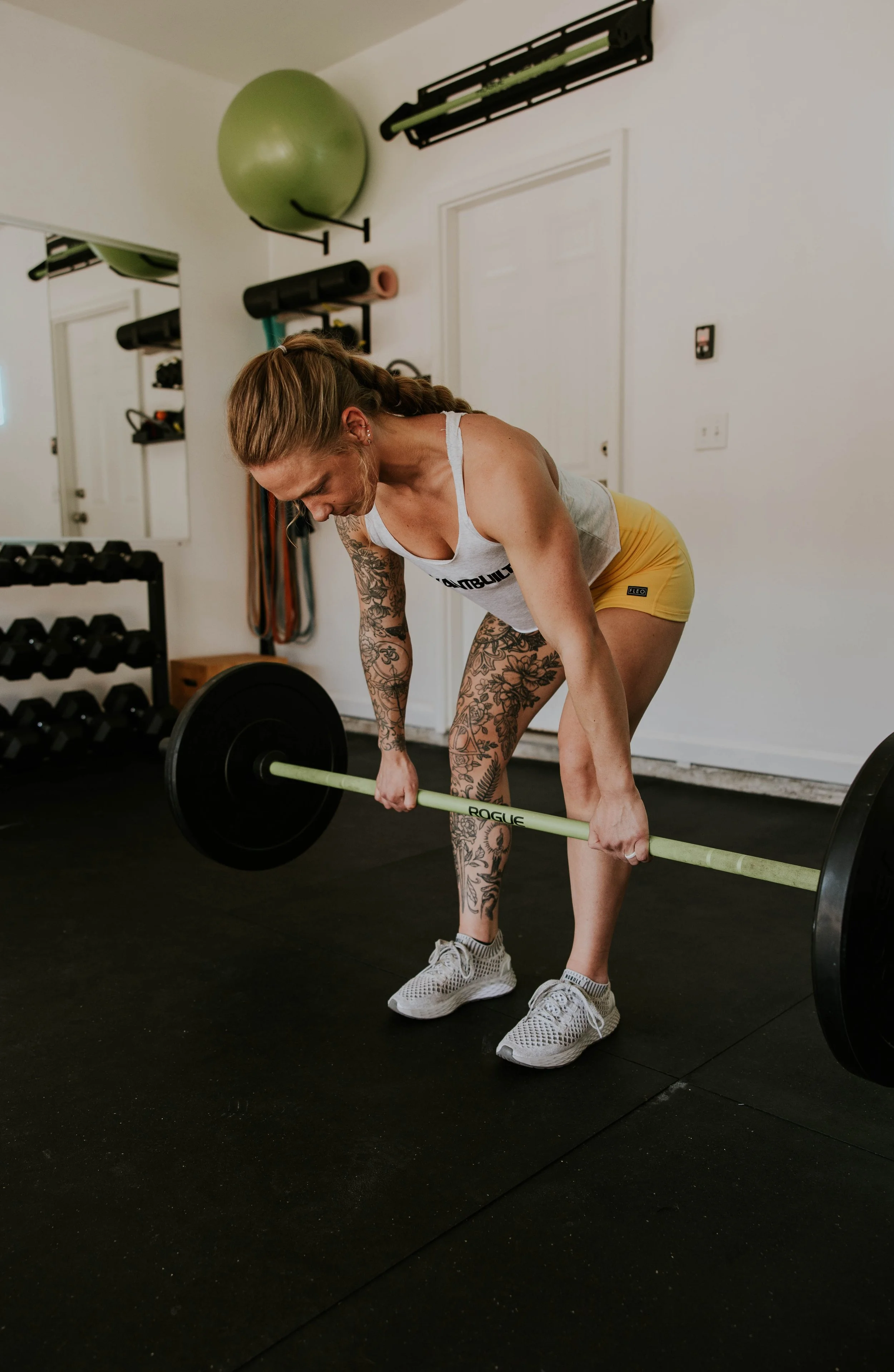 Woman in workout clothes bending over with a barbell on the ground in a home gym, surrounded by exercise equipment like a stability ball, dumbbells, and resistance bands.