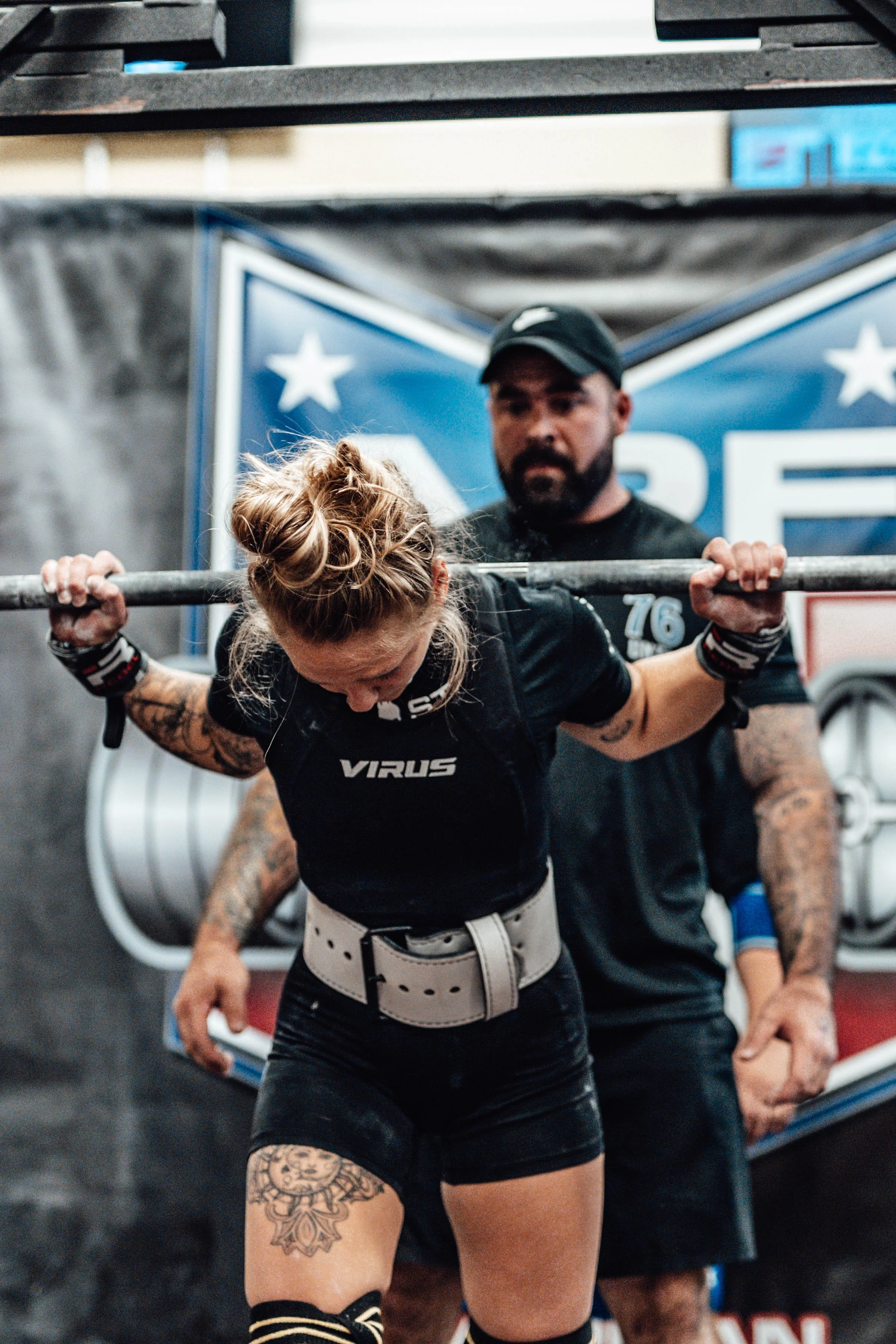 A woman doing a barbell squat exercise in a gym, with a trainer closely observing in the background, behind a blue and white star-themed banner.